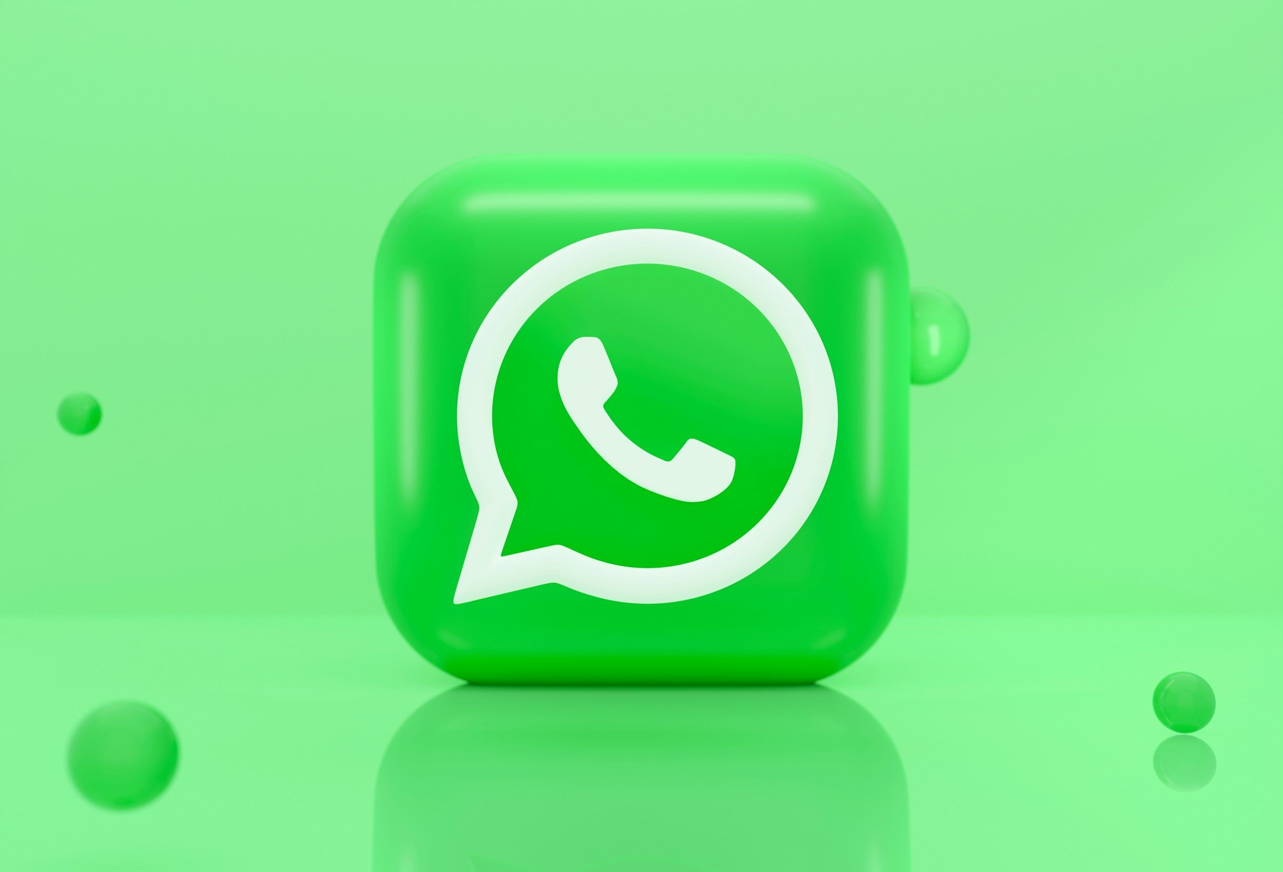 Close-up of a green cube with the white WhatsApp logo on its front, surrounded by small green and gray balls on a green background.