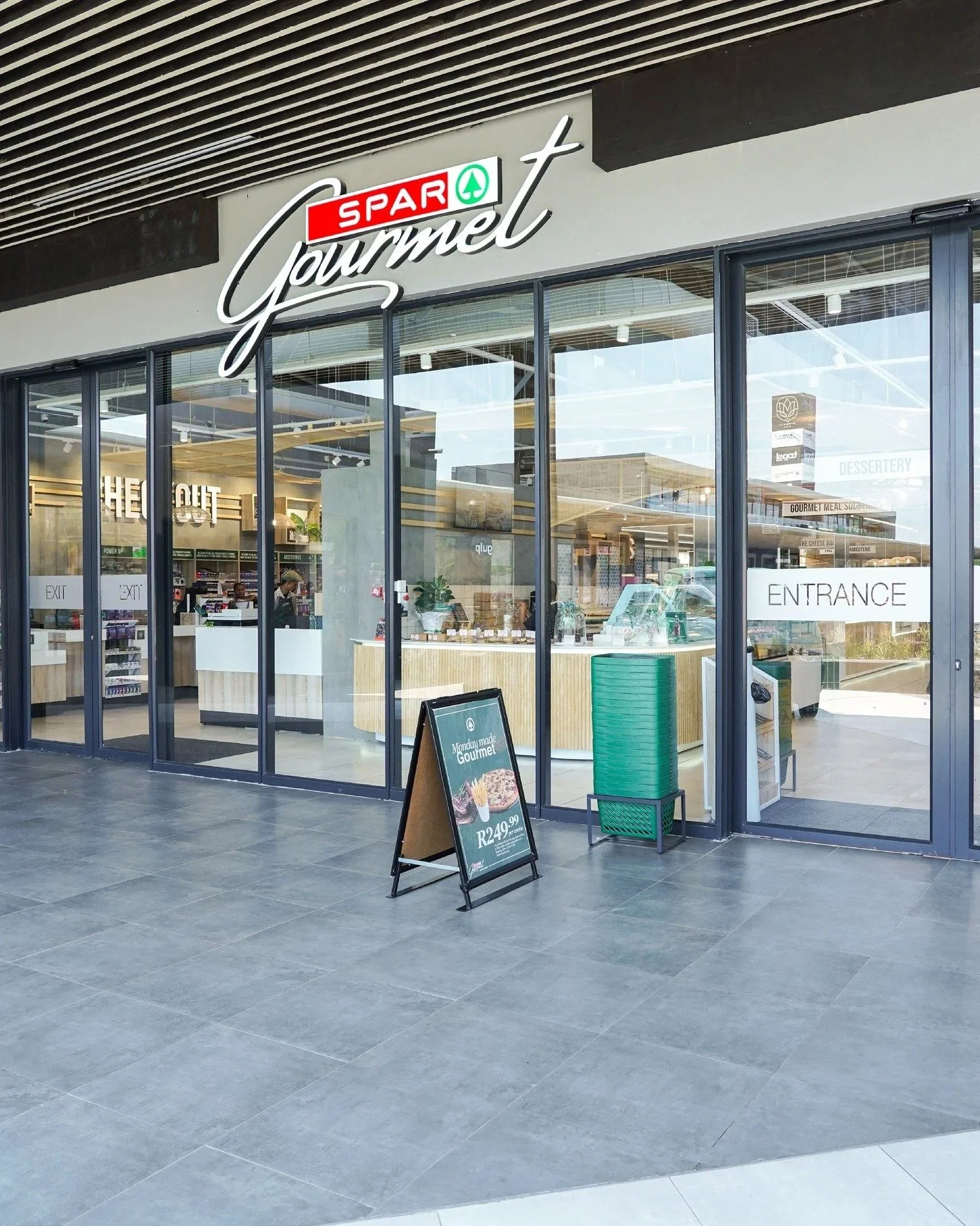 Exterior view of a grocery store named 'SPAR Gourmet' with large glass windows and doors. The store has a black sign with white cursive writing and a small red and green logo. There is a sandwich board sign outside the entrance advertising a gourmet 