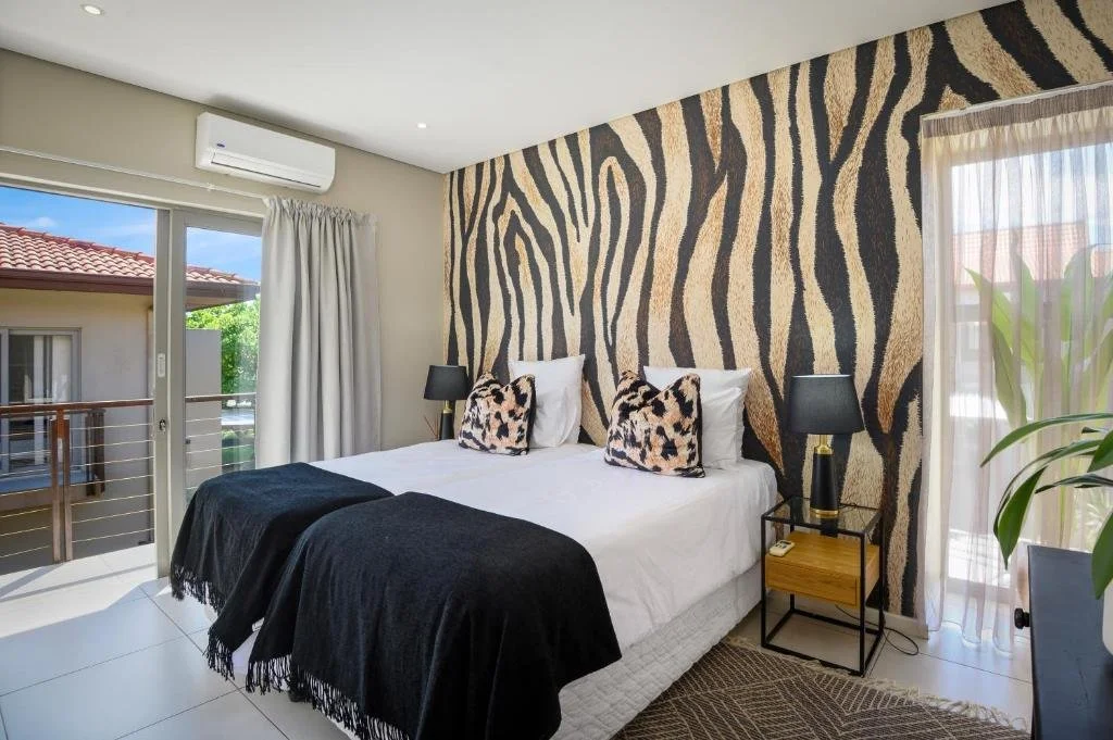 Bedroom with a large bed, animal print pillows, black and beige zebra print wallpaper, black lamps, and sliding glass doors with white curtains leading to a balcony.
