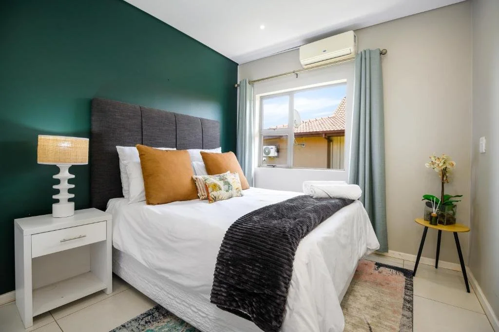 A bedroom with a bed, dark green accent wall, white nightstand with lamp, window with curtains, and various pillows on the bed.