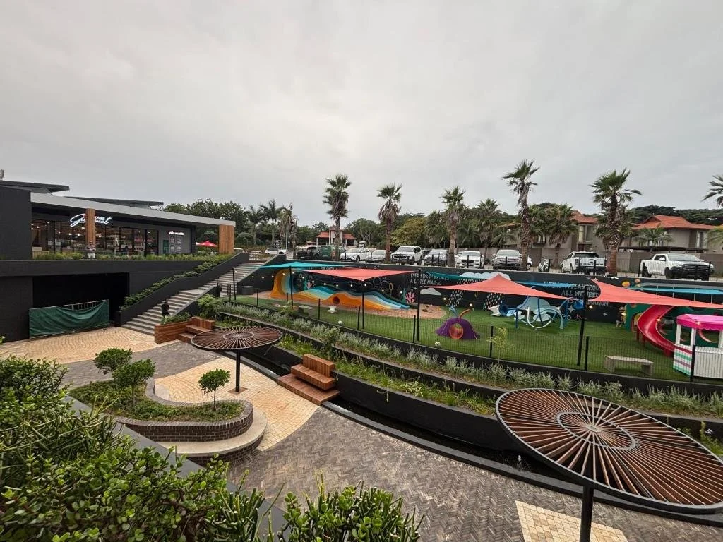 Outdoor area with colorful playground equipment, palm trees, a restaurant, and parking lot on a cloudy day.