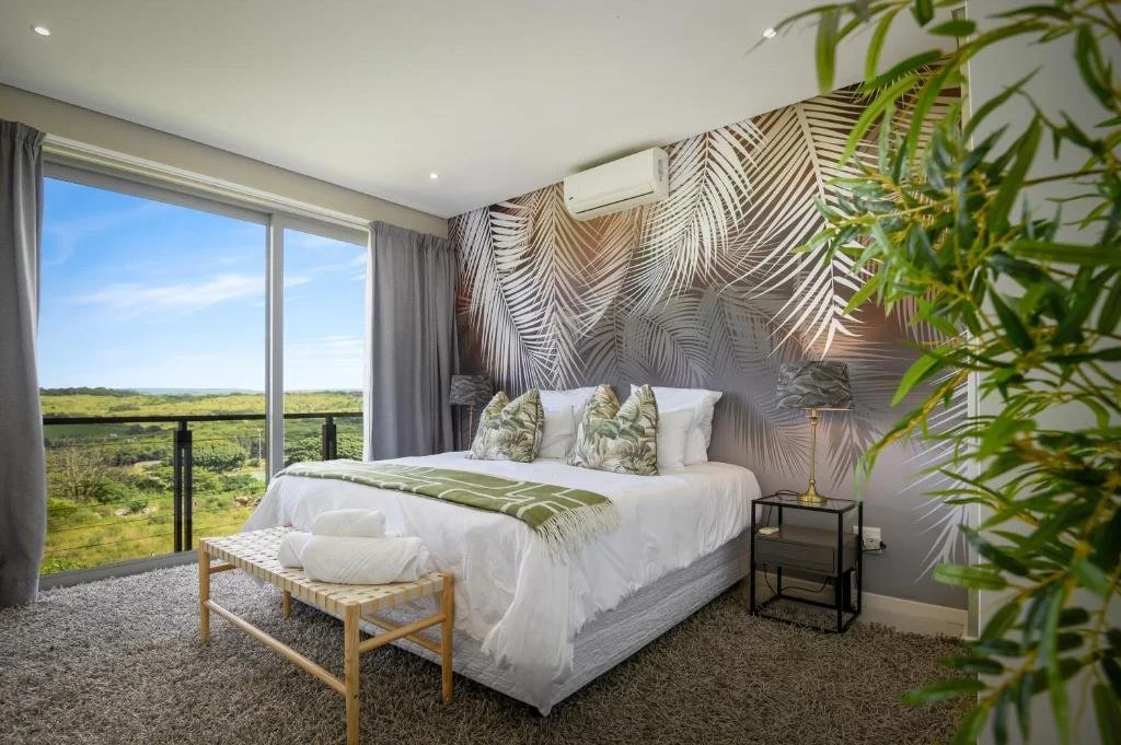 A bedroom with a bed, decorative pillows, a bench at the foot of the bed, a nightstand with a lamp, a wall with a tropical leaf mural, an air conditioning unit, large windows showing a green landscape and blue sky.