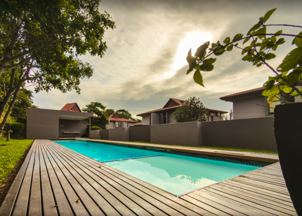 Residential backyard with a swimming pool, wooden deck, green grass, trees, and modern houses under a cloudy sky with the sun partially visible.