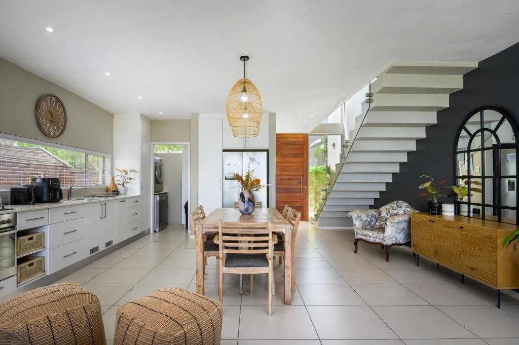 Open-concept kitchen and dining area with white cabinets, a wooden dining table with matching chairs, a clock on the wall, a black accent wall with a staircase, and a vintage armchair.