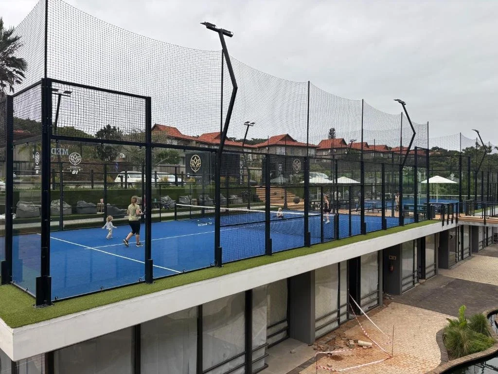 Outdoor padel tennis court with players and children, surrounded by a high net and fence, with umbrellas and residential buildings in the background.