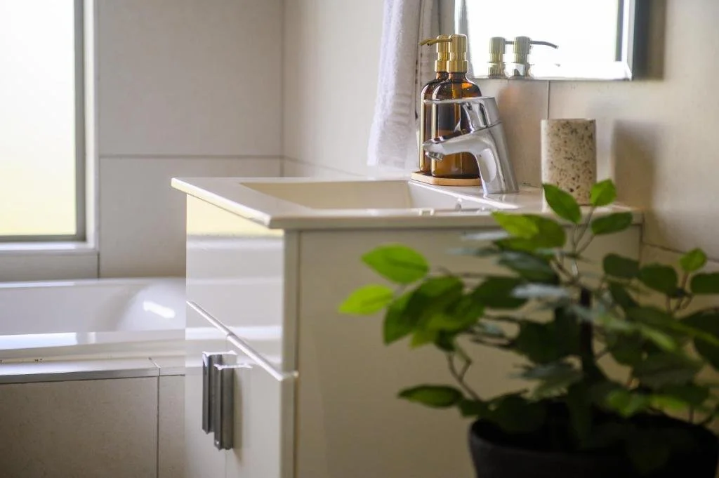 A bathroom sink with a gold soap dispenser, a clear soap pump, and a soap bowl on the counter. There is a window with white curtains, a potted green plant in the foreground, and a bathtub partially visible.