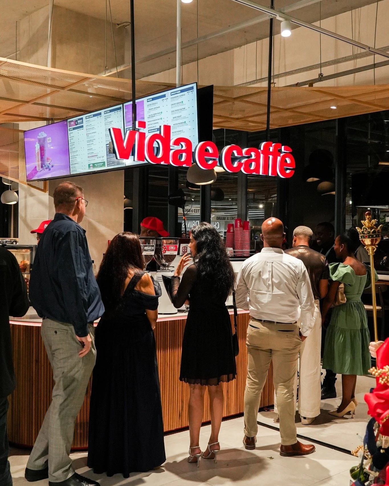 People standing in line at a cafe counter with a red sign that says 'video café' above. The cafe has a digital menu screen and a modern interior design.