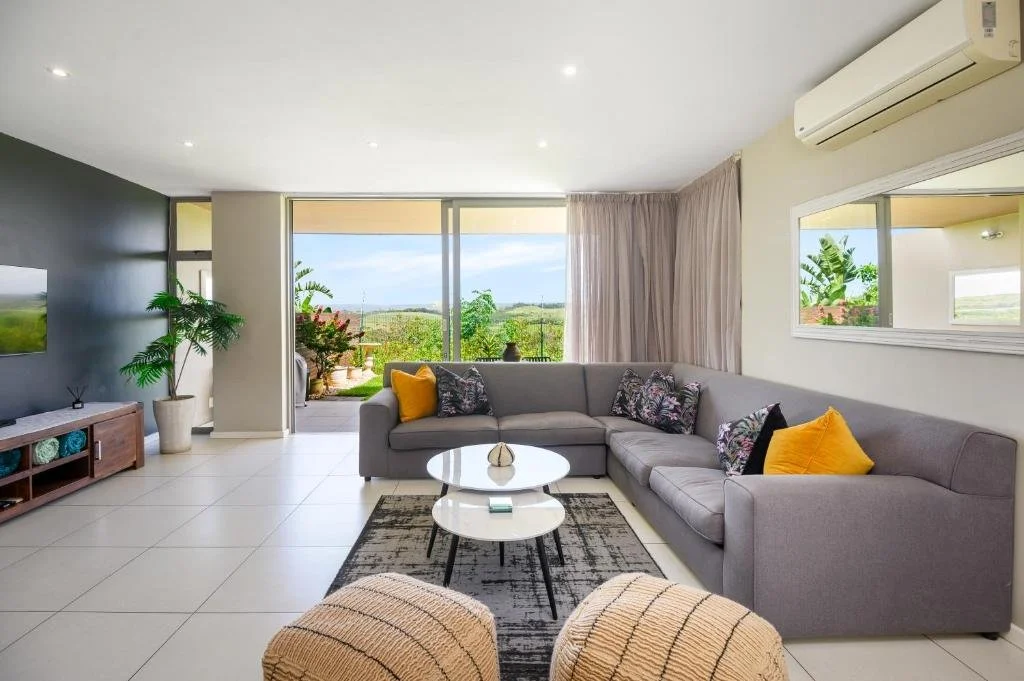 Modern living room with gray sectional sofa, colorful throw pillows, glass sliding door leading to outdoor patio with green landscape, white tiled floor, TV, potted plant, and large mirror on cream wall.