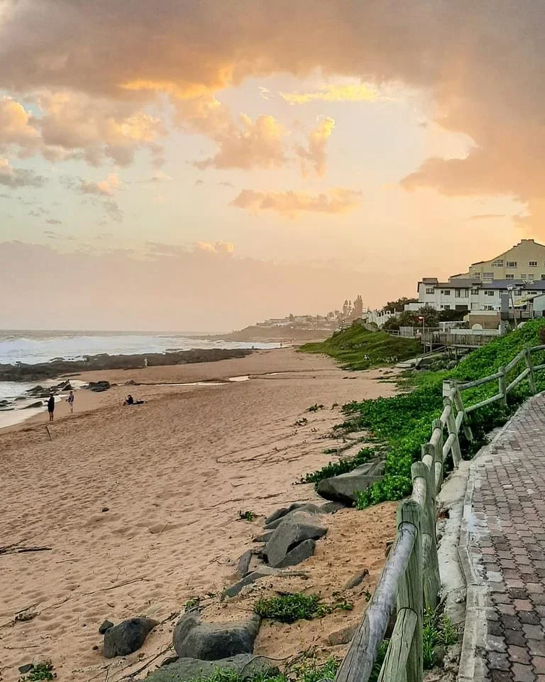 A sandy beach with a walkway and railing on the right side, some rocks and green plants near the shore, houses on a hill in the distance, and a partly cloudy sky with warm sunset colors.