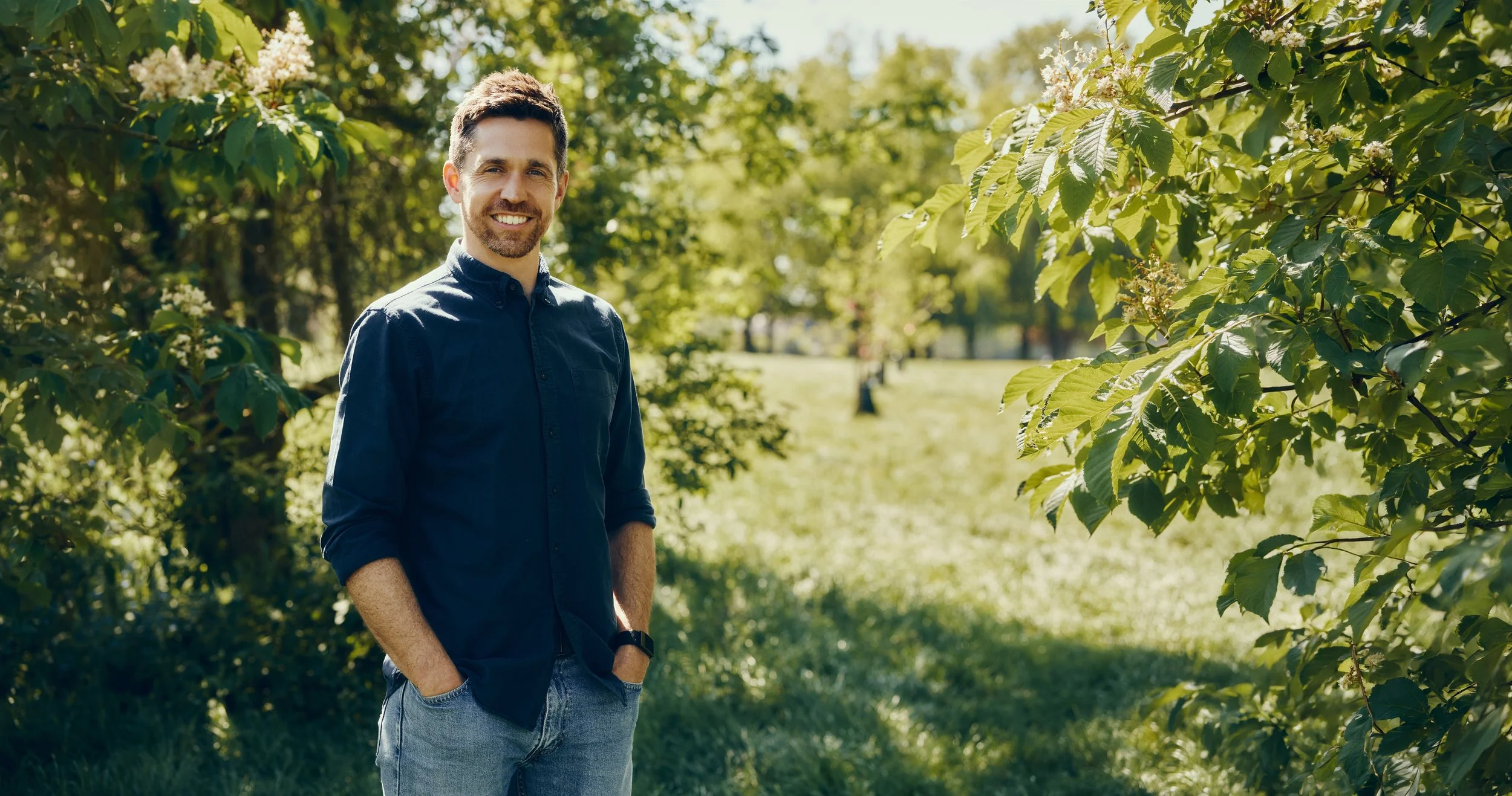 A man with a beard and short dark hair standing outdoors among green trees, smiling, wearing a dark blue shirt and jeans, with hands in pockets on a sunny day.