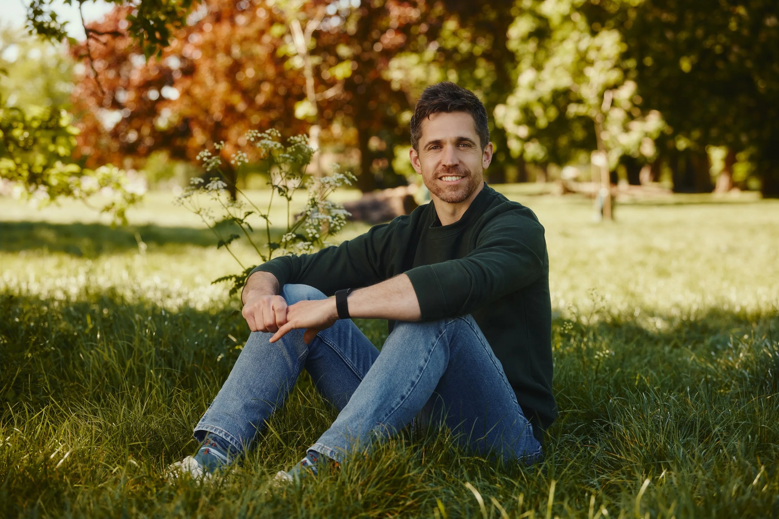 A man sitting on the grass in a park with trees and autumn foliage in the background, smiling and looking at the camera.