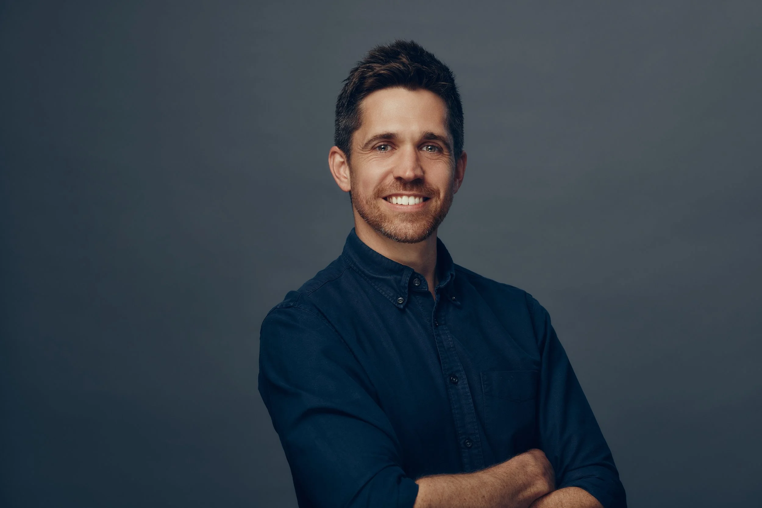 Portrait of a smiling man with dark hair and a beard, wearing a dark blue button-up shirt, against a gray background.