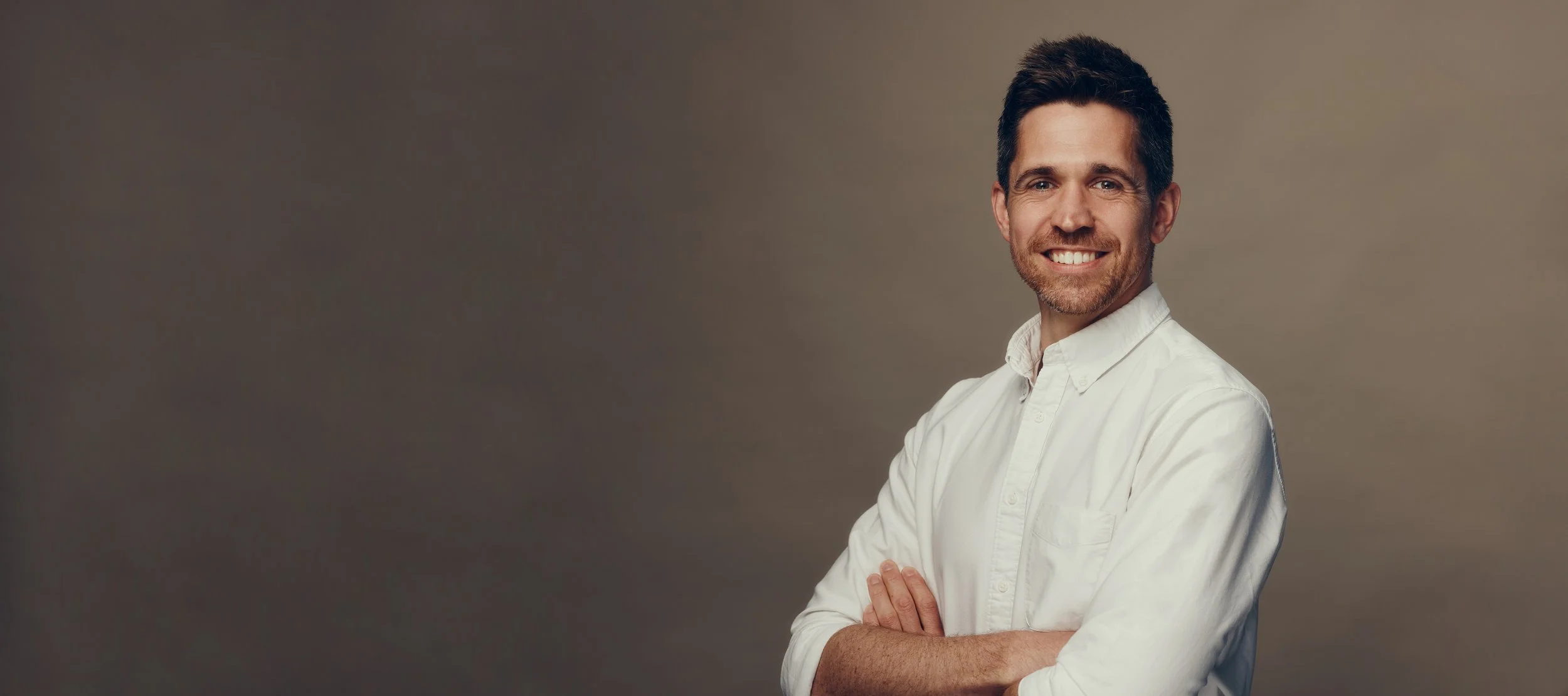 A man with dark hair and a beard wearing a white shirt, smiling with arms crossed, against a neutral background.