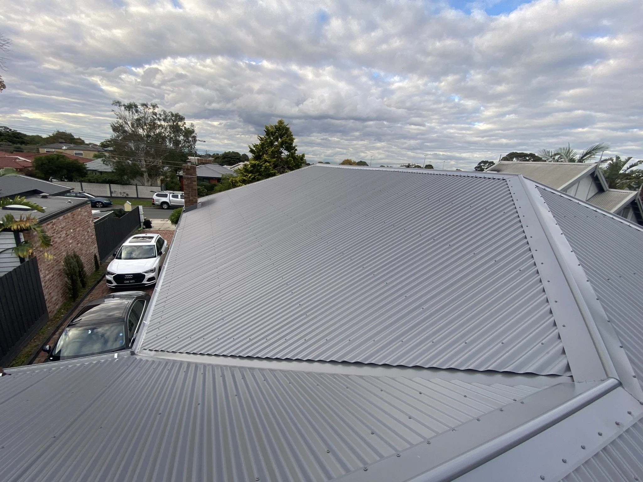 View of rooftops with grey metal roofing, parked cars, trees, and a cloudy sky.