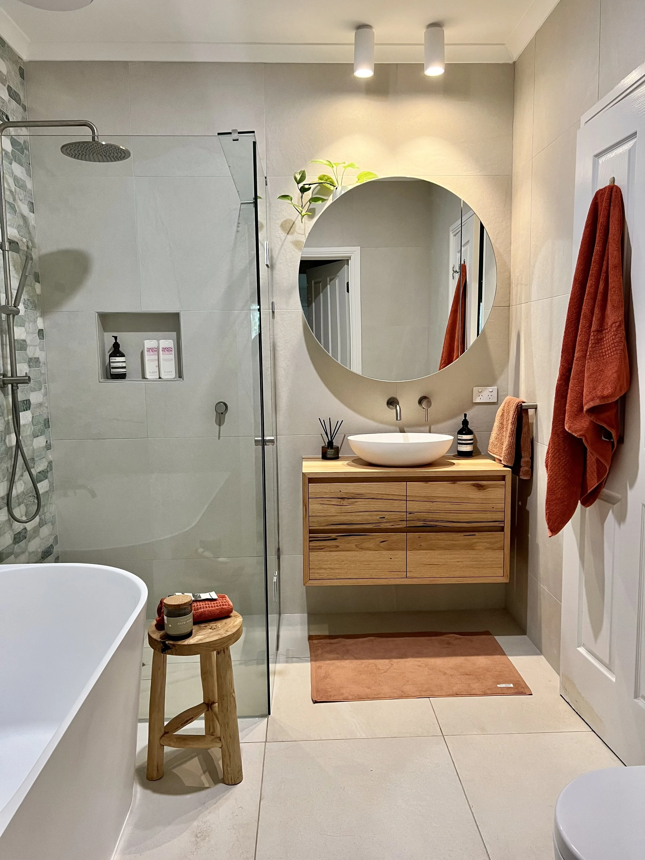 Modern bathroom interior with a walk-in shower, wooden vanity with a vessel sink, round mirror, orange towels, and beige tiles.