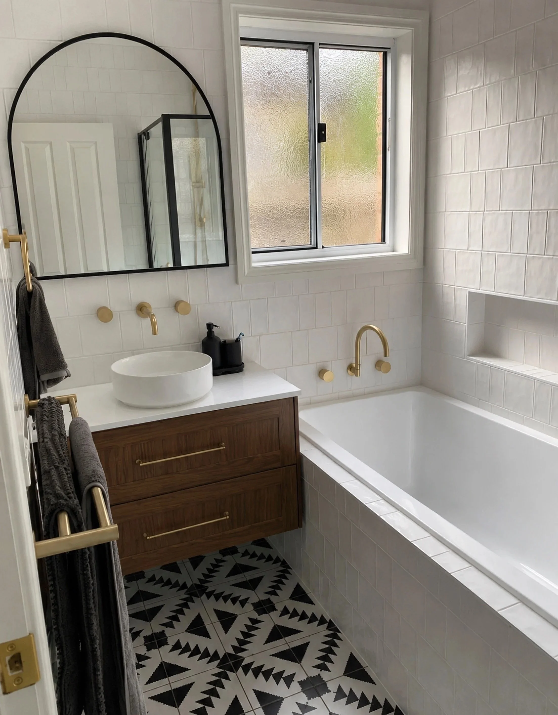 Bathroom with large window, black and white geometric patterned floor tiles, white brick walls, a wooden vanity with a white vessel sink, black faucets, gold fixtures, and a mirror reflected in the window showing a shower with a black frame.