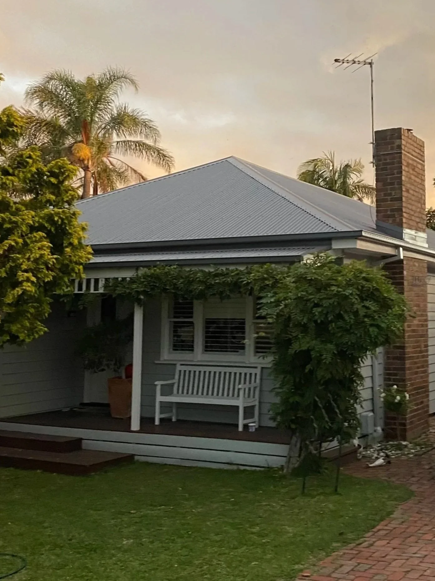 A house with a gray corrugated metal roof, a brick chimney, and an outdoor porch with white furniture, surrounded by trees and greenery, with an overcast sky.