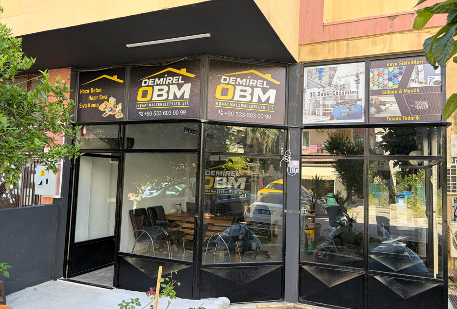 Bir inşaat malzemeleri dükkanının önü, siyah çerçeveli büyük cam pencereleri ve kapısı var. Üstte, Demirel OM isimli inşaat ve yapı malzemeleri dükkanına ait ilanlar bulunuyor. Dükkanın tabelasında çeşitli inşaat malzemeleri ve teknik çözümler ile ilgili bilgiler var.