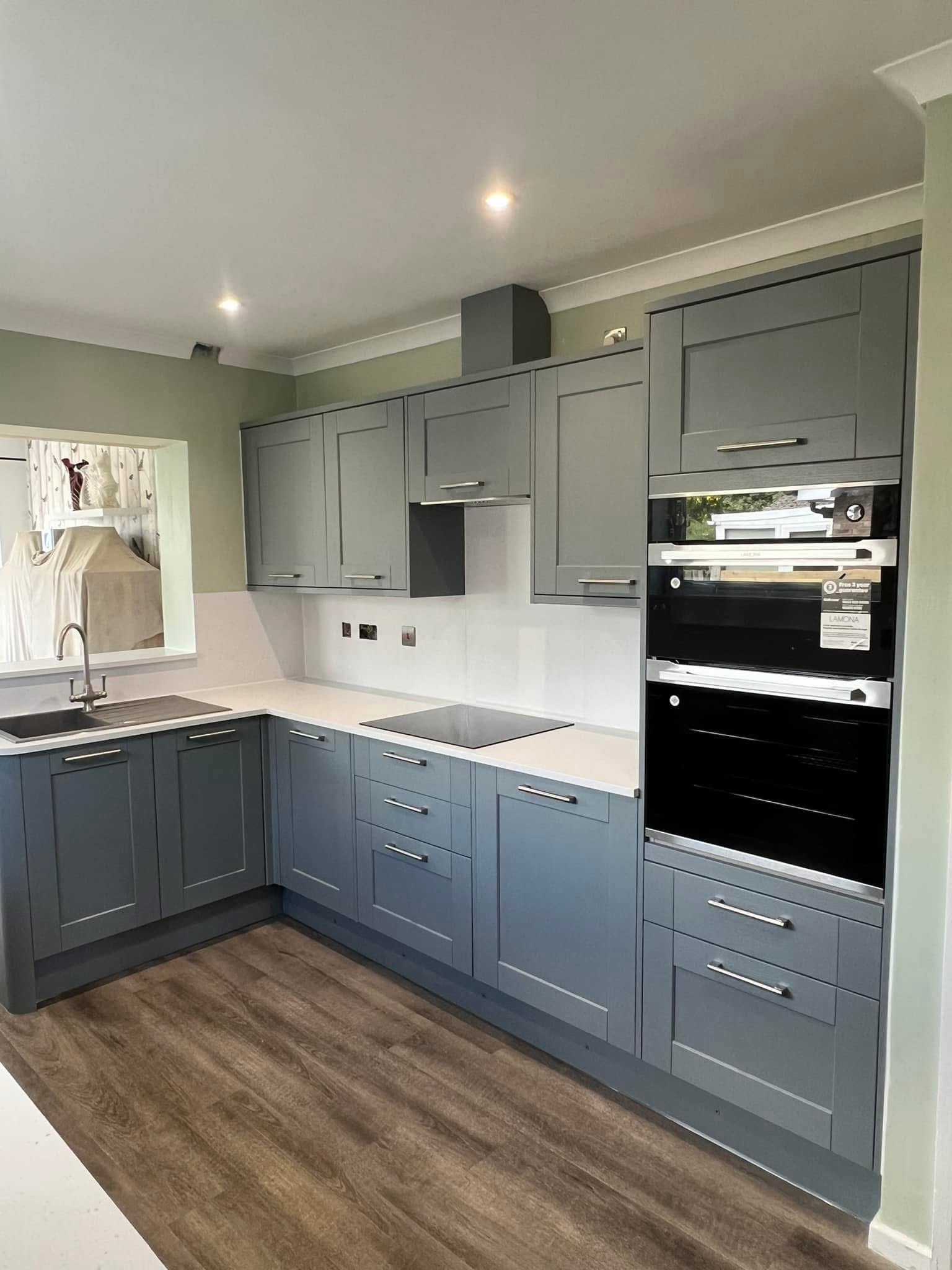 Modern kitchen with gray cabinets, a white countertop, a built-in oven, a stovetop, and a sink with a faucet. There is a window above the sink, and wooden flooring.