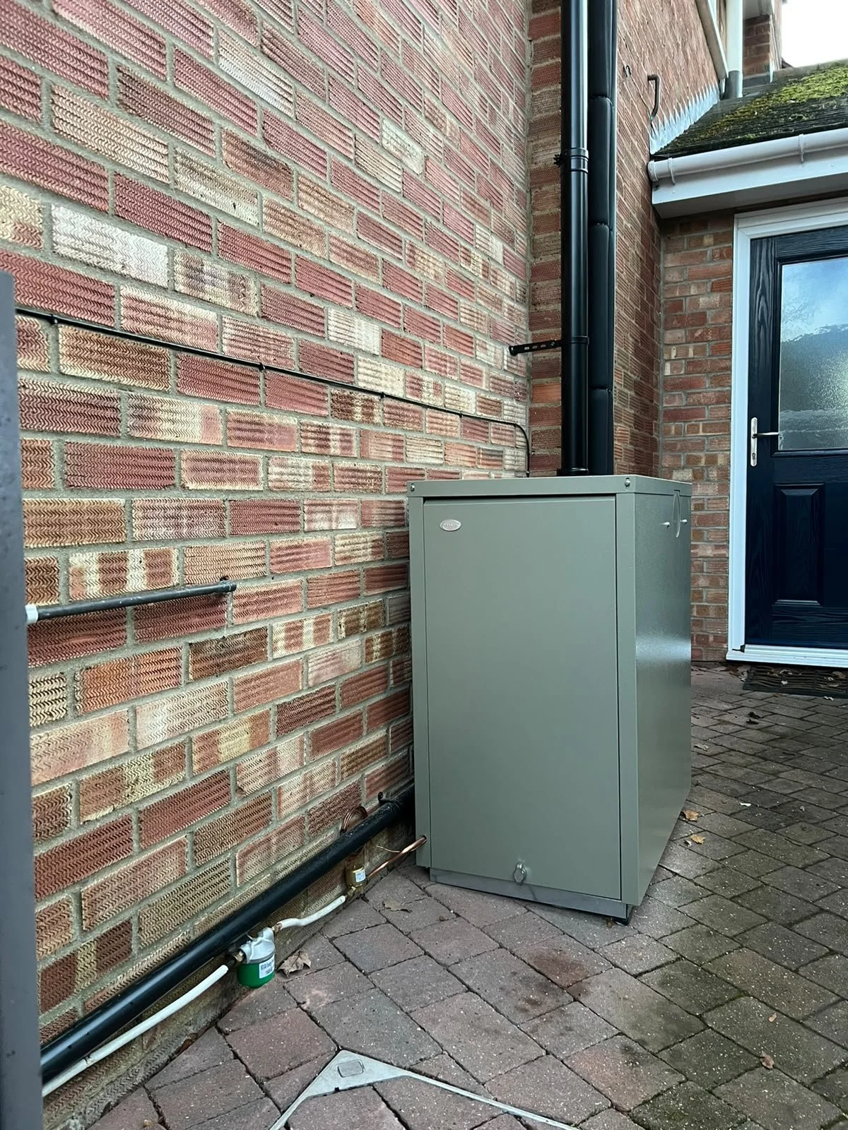 Black outdoor door next to a brick wall with a black pipe and a utility box on the ground outside.