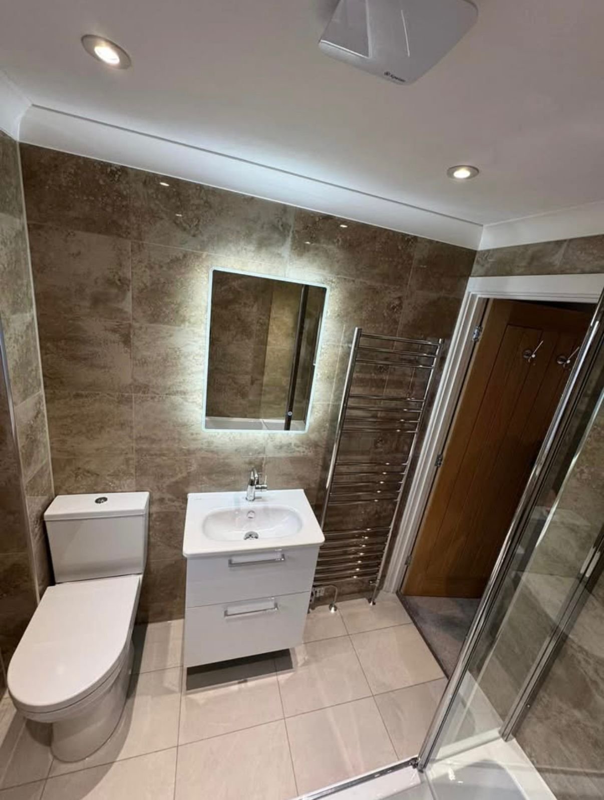 A modern bathroom with beige tiled walls and floors, a white ceramic toilet, white sink with vanity, a wall-mounted mirror with backlighting, a metal towel radiator, a wooden door, and a glass shower enclosure.