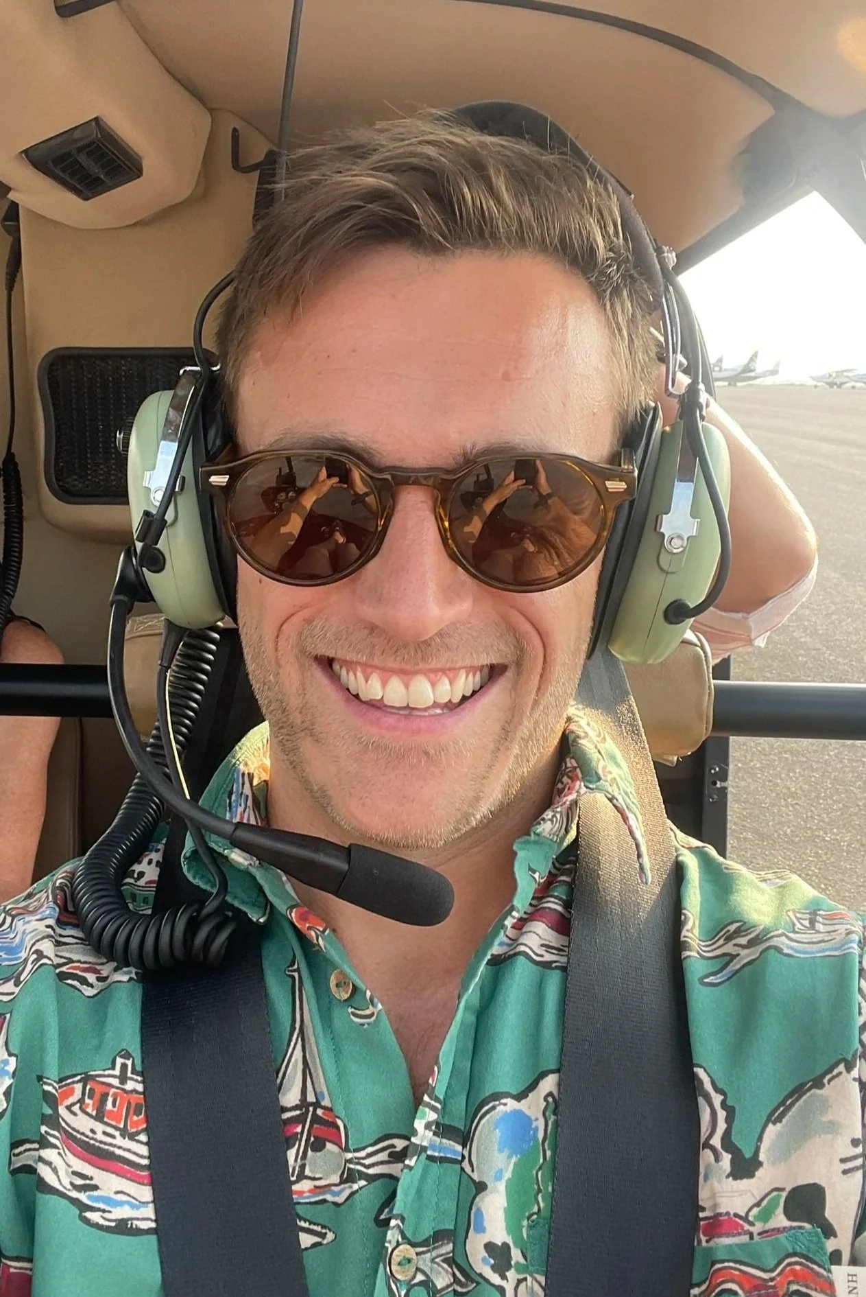 Hugo Morrissey, wearing sunglasses and a green shirt with a colorful pattern, sitting in a helicopter with a headset and microphone, with airport runway and airplanes visible in the background.