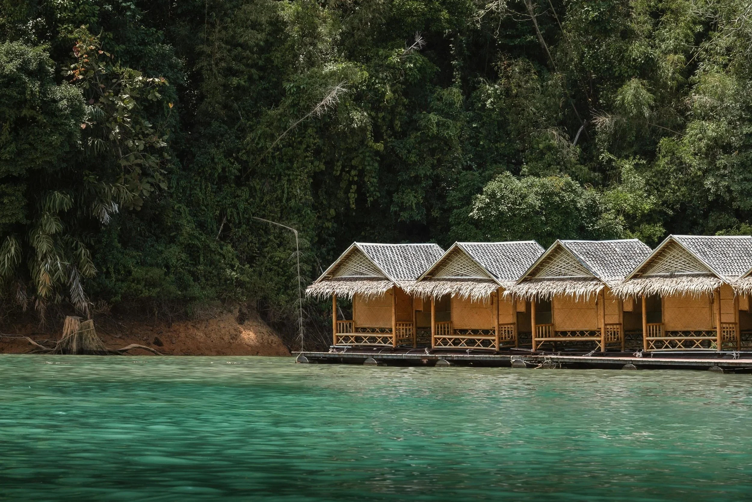 Overwater thatched-roof huts on stilts over turquoise water in Indonesia, with lush tropical greenery on the shoreline in the background.