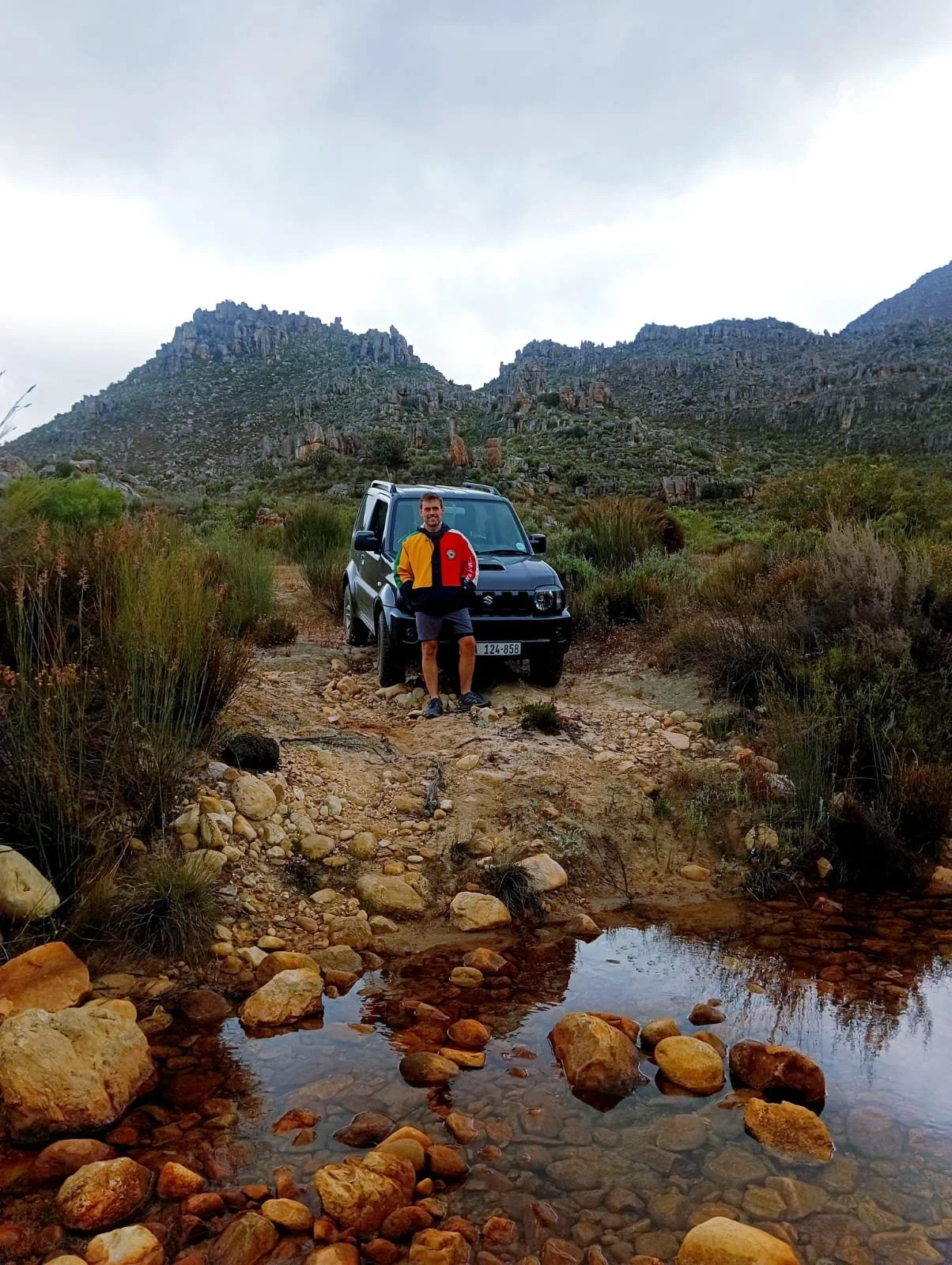Hugo Morrissey, founder, standing in front of a black Suzuki Jimny vehicle on a rocky dirt trail in a mountainous landscape with sparse vegetation. Gray clouds are overhead, and there is a small water pool with rocks in the foreground.