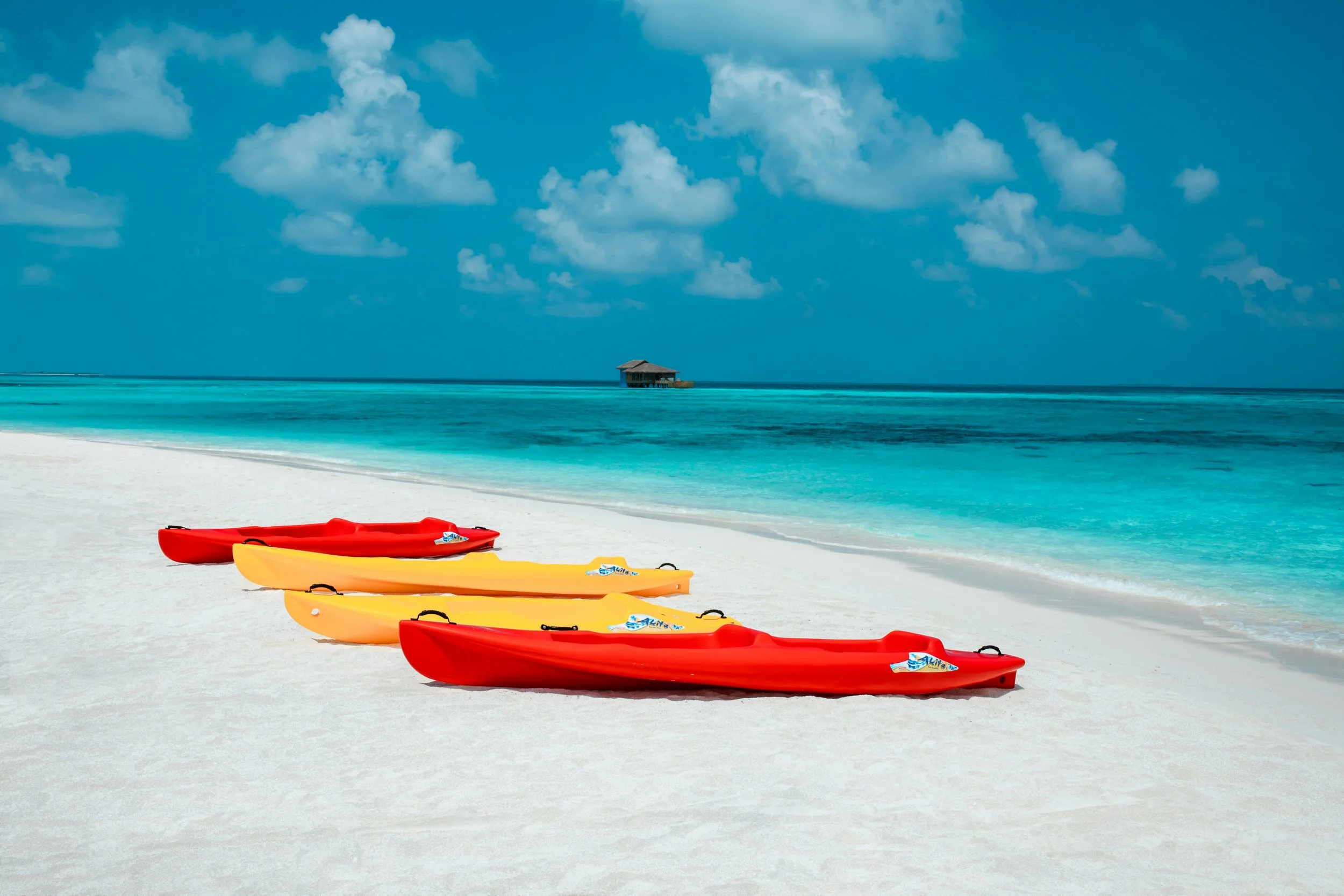 Colorful kayaks on a white sandy Maldives beach with turquoise water and a small hut on stilts in the distance, under a partly cloudy sky.