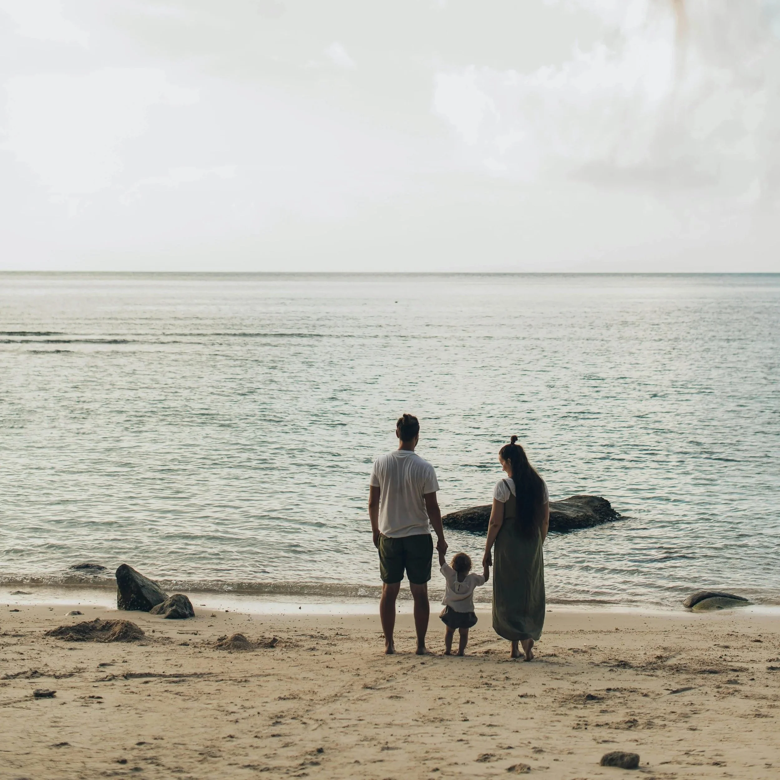 A family of three, including a man, a woman, and a young child, walks hand in hand along the Cape Town beach at sunset. The man and woman are near large rocks at the shoreline, with the ocean and cloudy sky in the background.