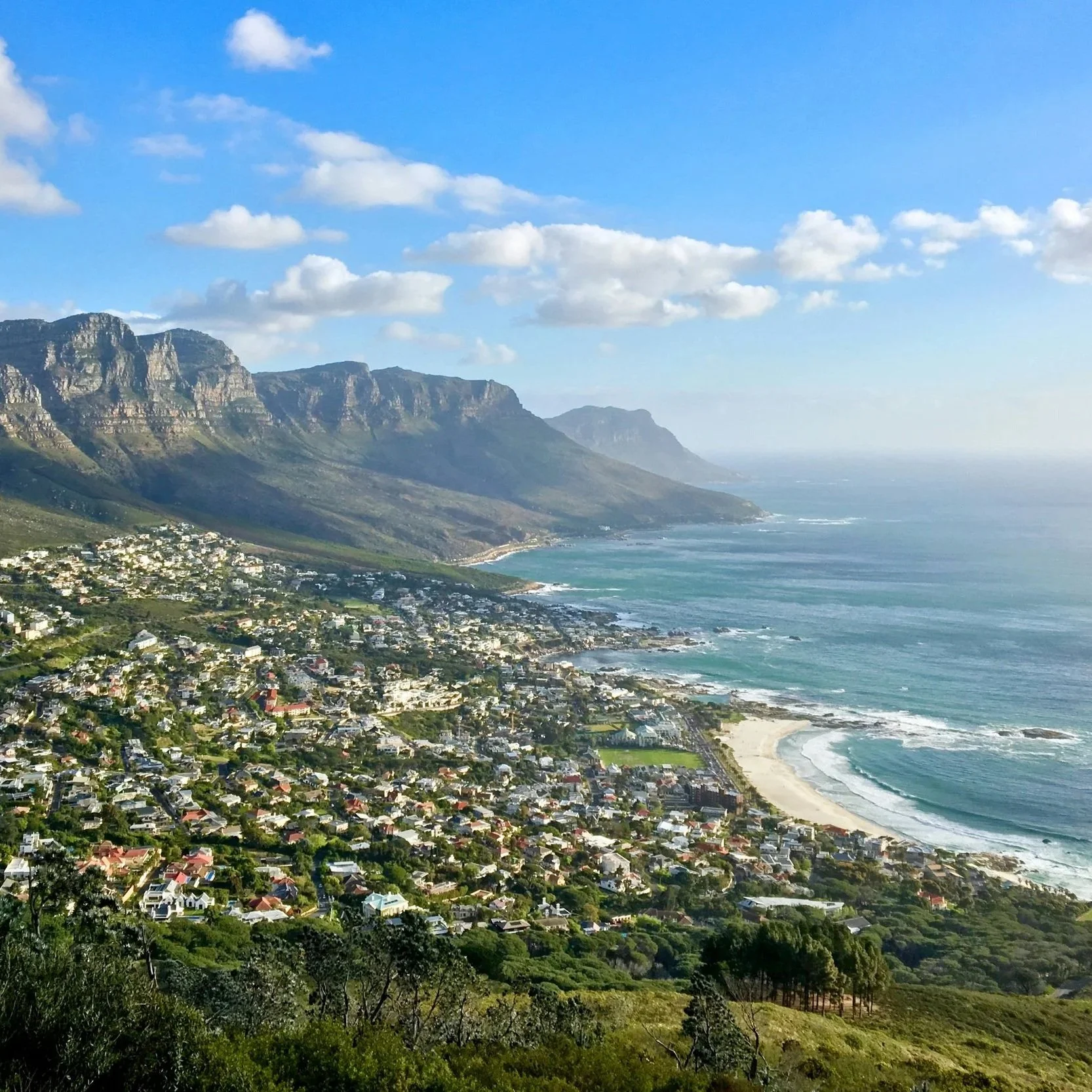 Cape Town Camps Bay view with houses along the shoreline, green hills in the foreground, mountain cliffs in the background, and a blue sky with clouds.