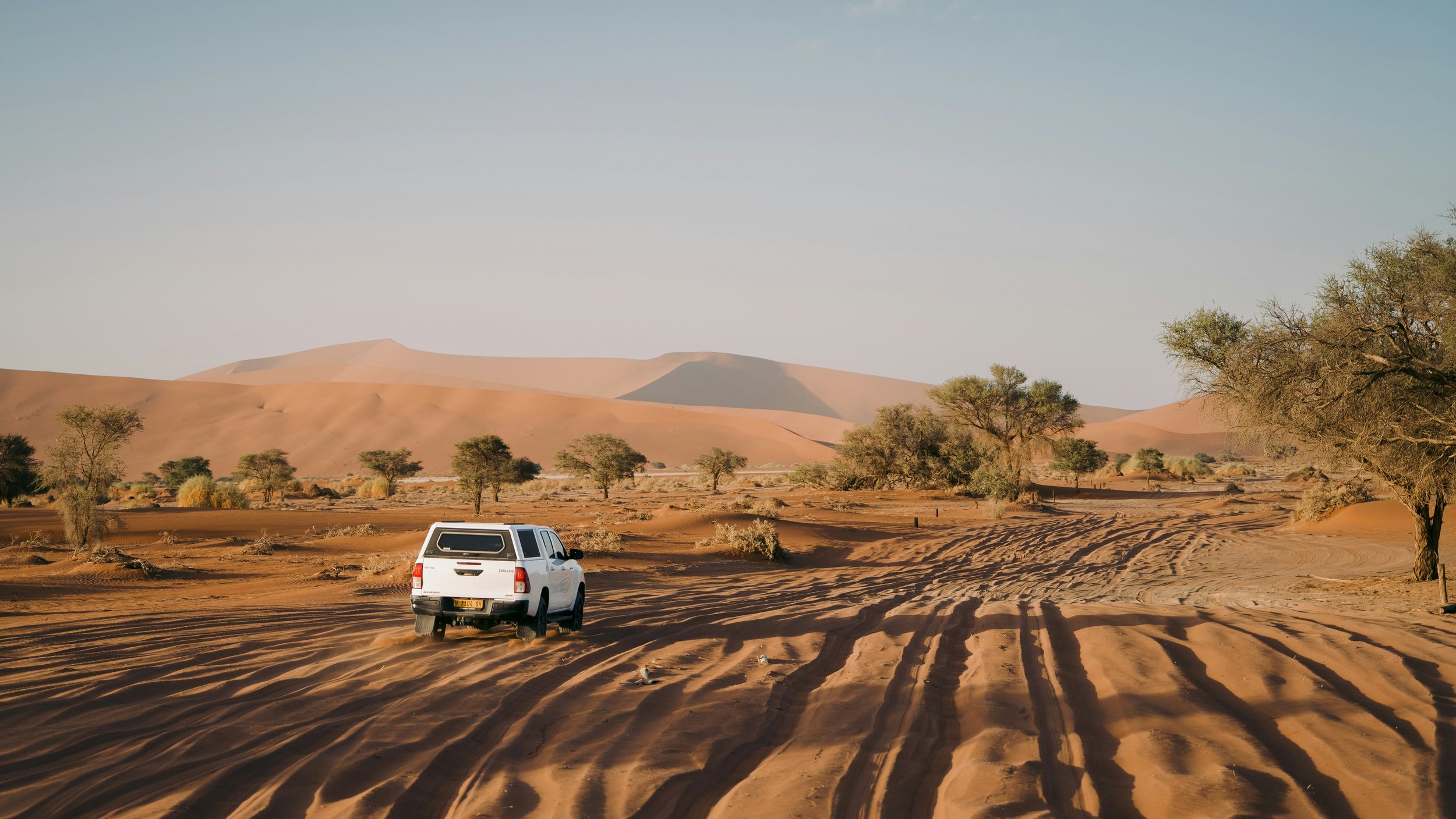 A white vehicle parked on sand dunes in an African desert landscape with scattered trees and distant sand dunes under a clear sky.