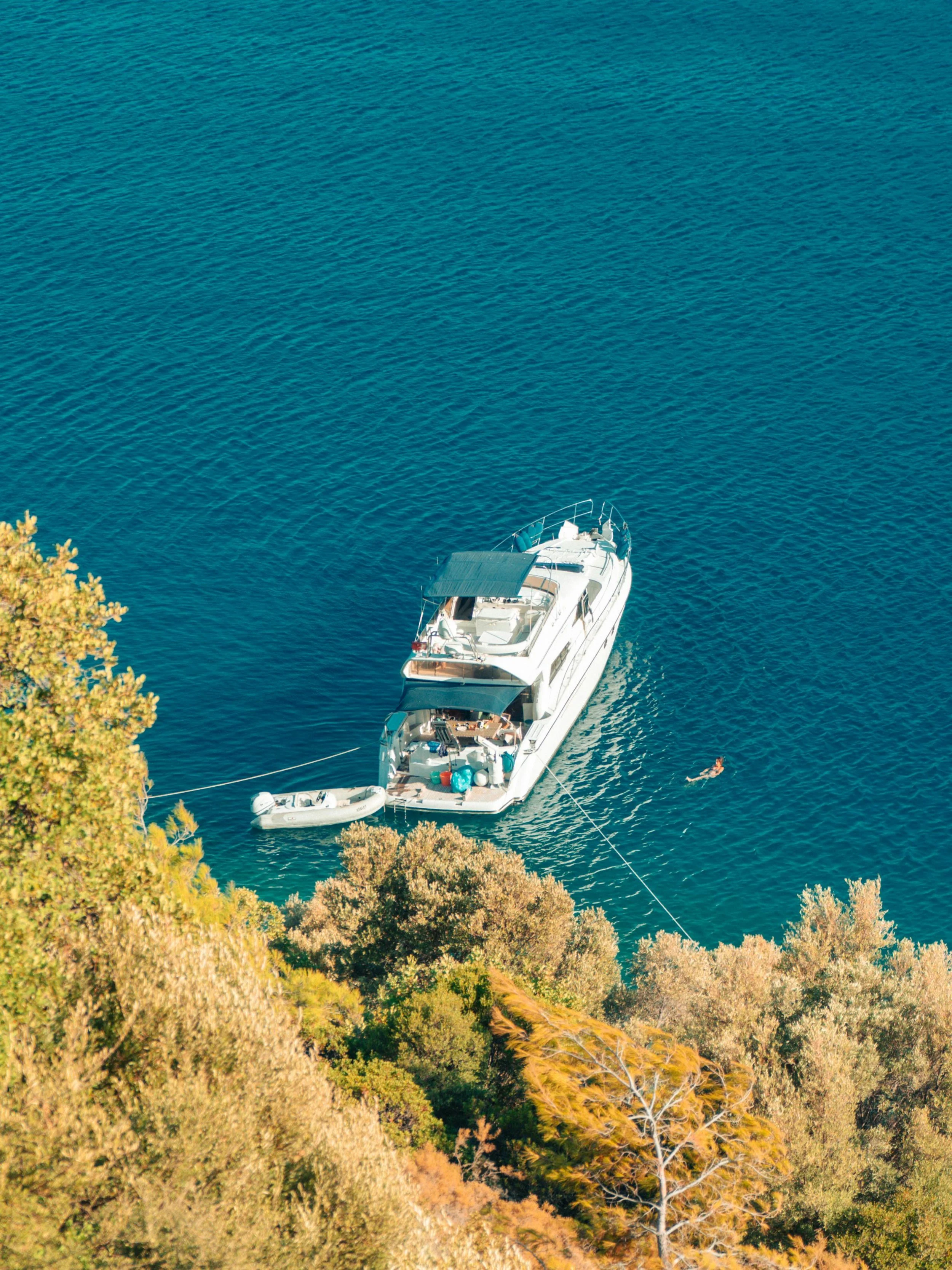A large yacht anchored near the green, orange, and yellow trees along a shoreline with blue water in the Mediterranean.