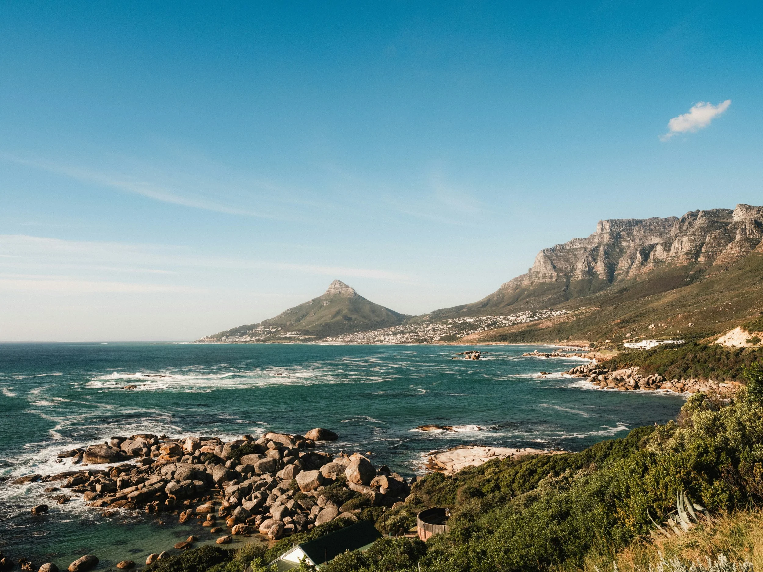 Cape Town in all it's glory. A coastal landscape with rocky shoreline, green vegetation, Lion's Head and Table Mountain in the background, and a partly cloudy sky.