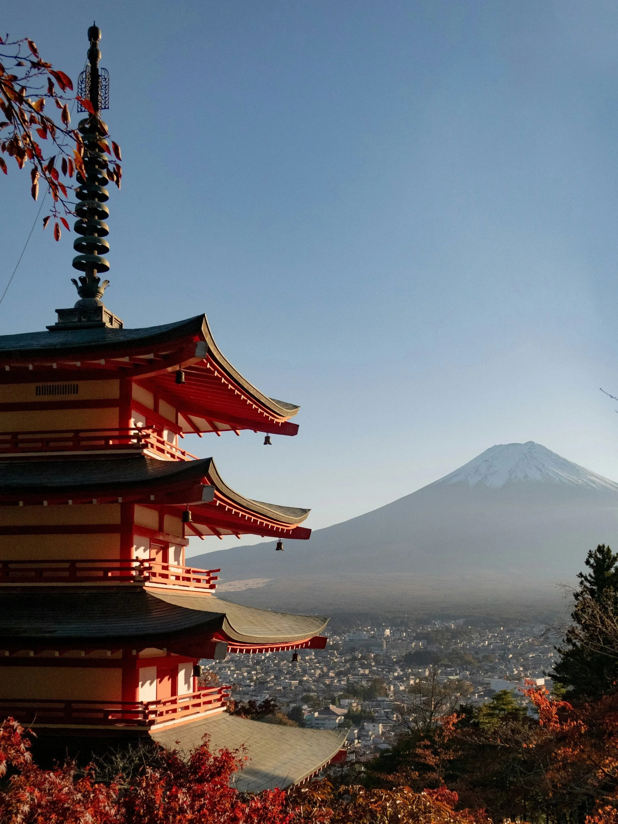 A traditional Japanese pagoda with multiple tiers and red accents, overlooking a cityscape with Mount Fuji in the background, under a clear blue sky.