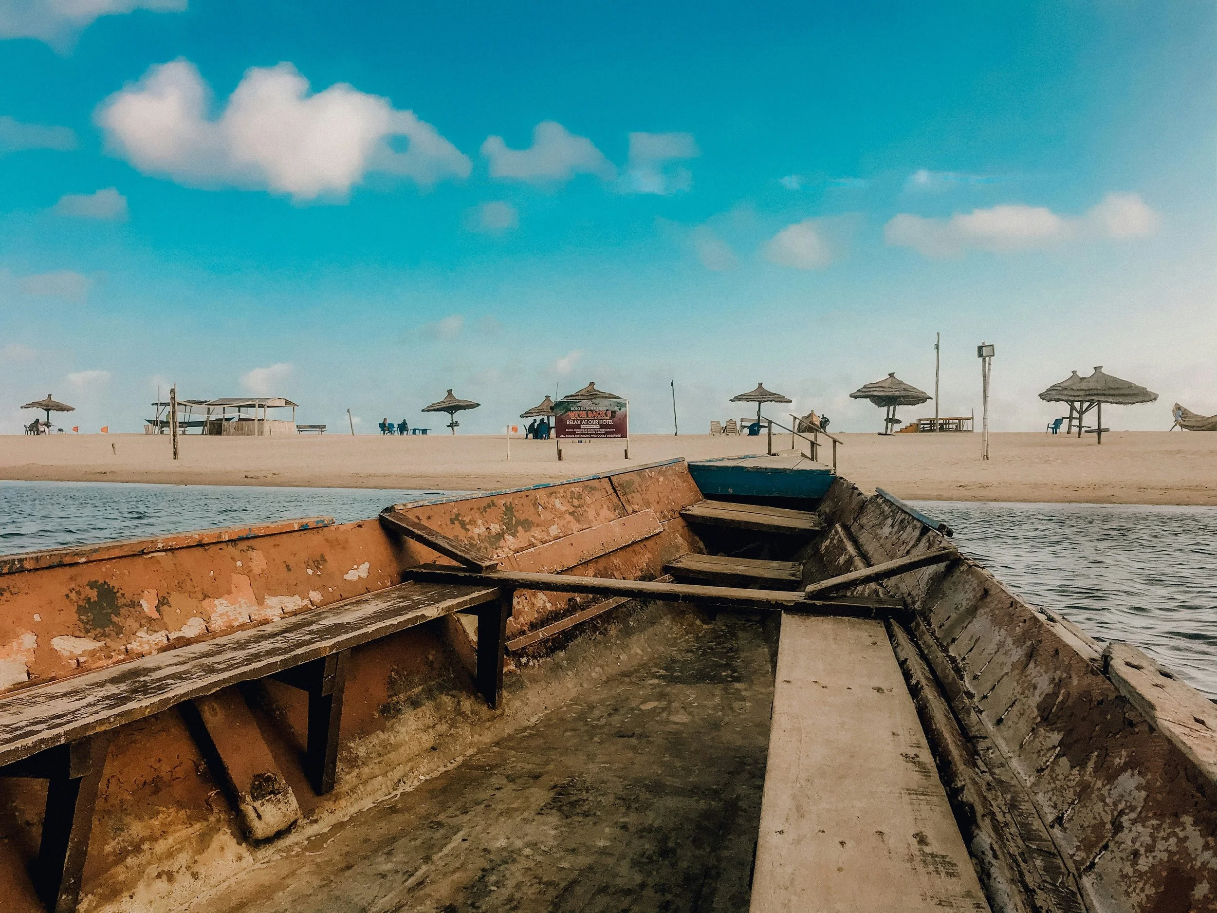 Rusty old boat docked at a beach in Mozambique with wooden steps and a distant sand beach with parasols and chairs under blue sky with clouds.