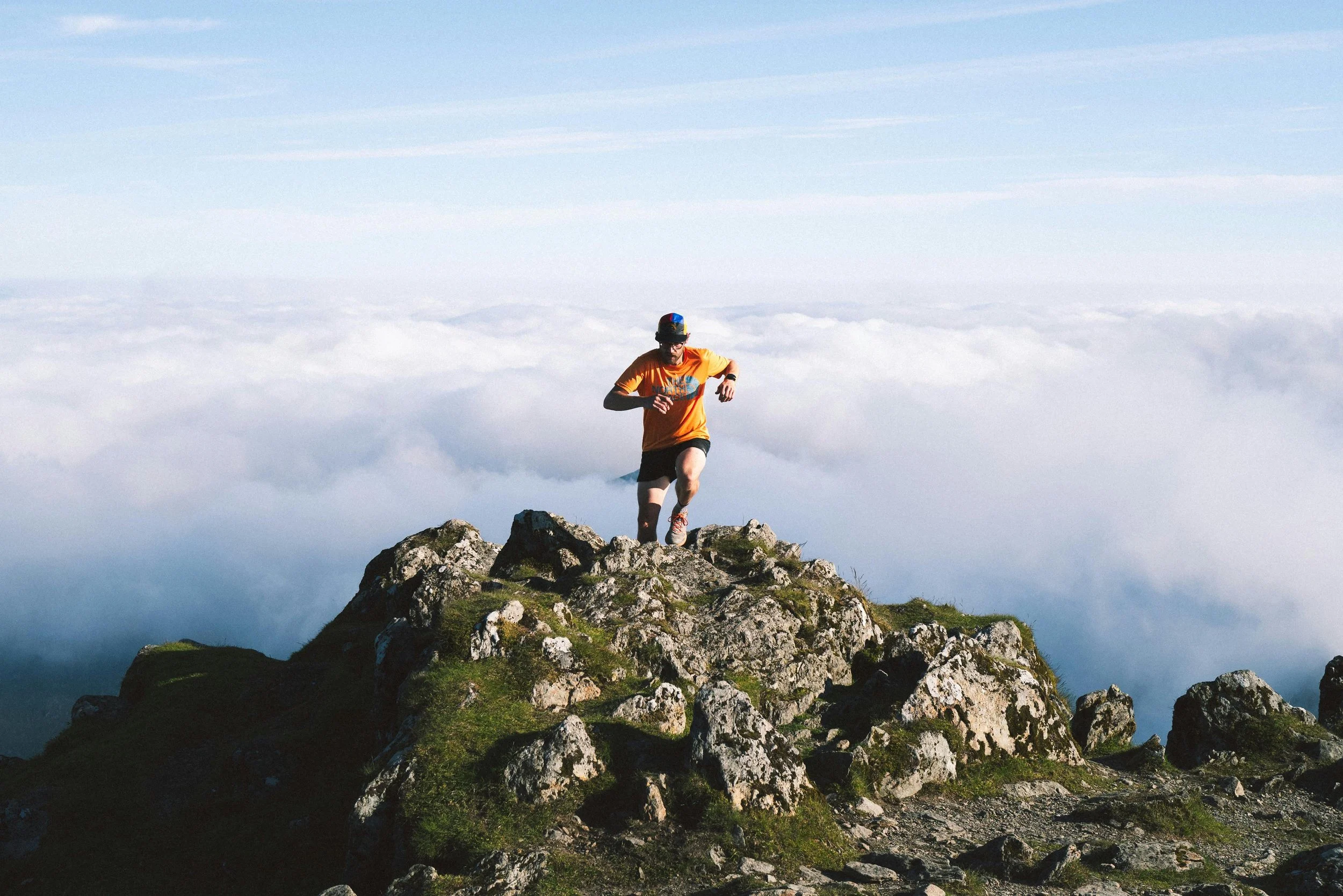 A man running on rocky South Africa mountain terrain above the clouds, wearing an orange T-shirt, black shorts, and a blue and black cap.