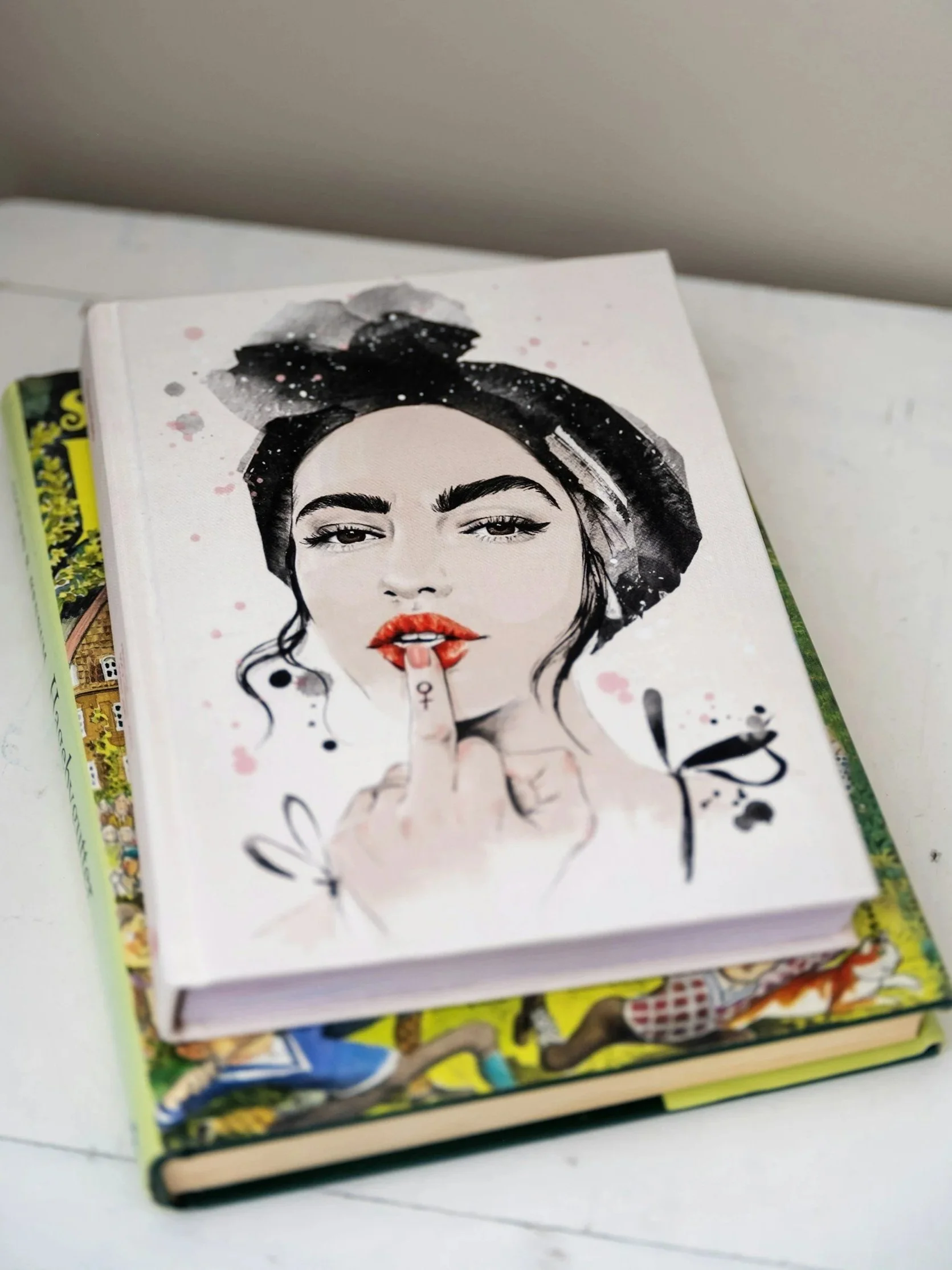 A book with an illustrated cover featuring a woman with dark hair, red lips, and her finger pressed against her lips, surrounded by abstract paint splashes.