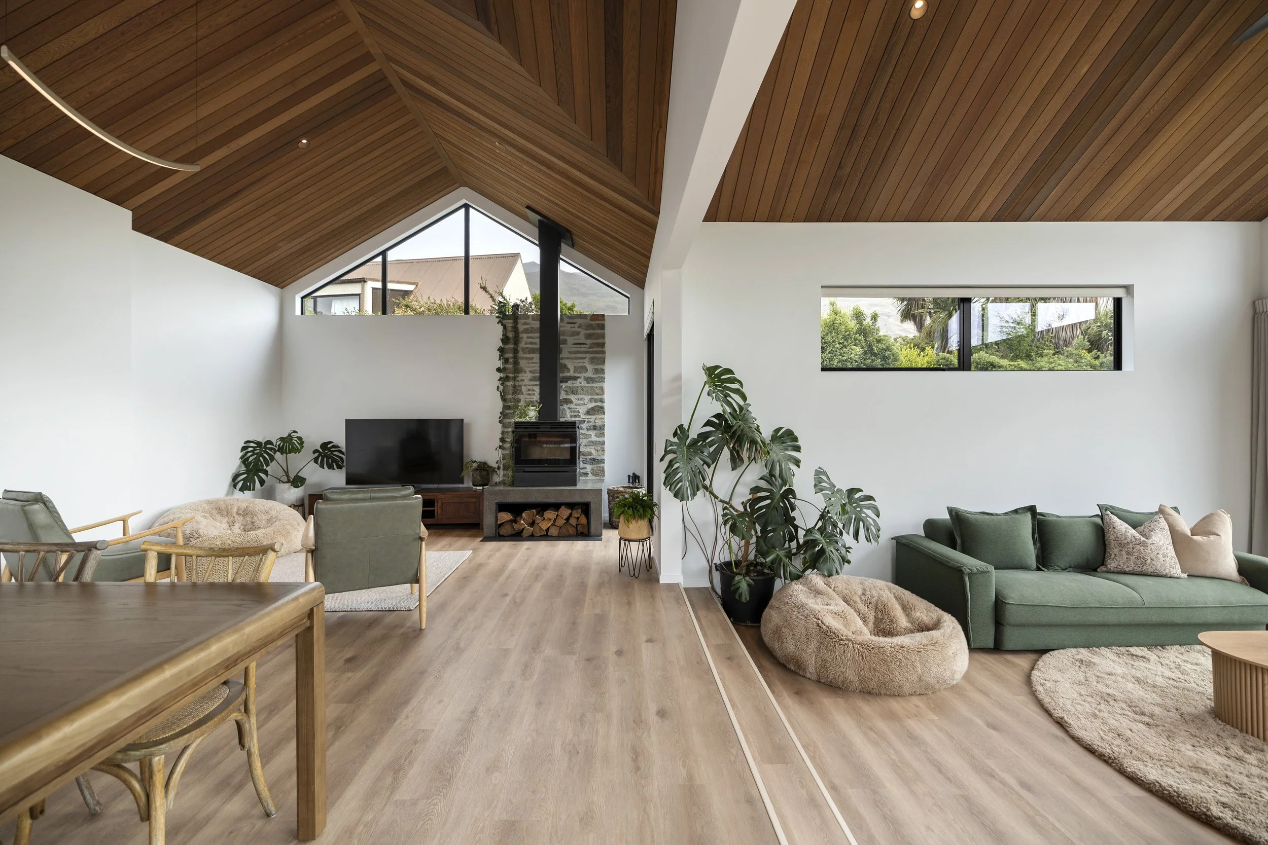Modern living room with wooden vaulted ceiling, white walls, large windows showing greenery outside, green sofa with pillows, beige bean bag, indoor plants, and a fireplace.