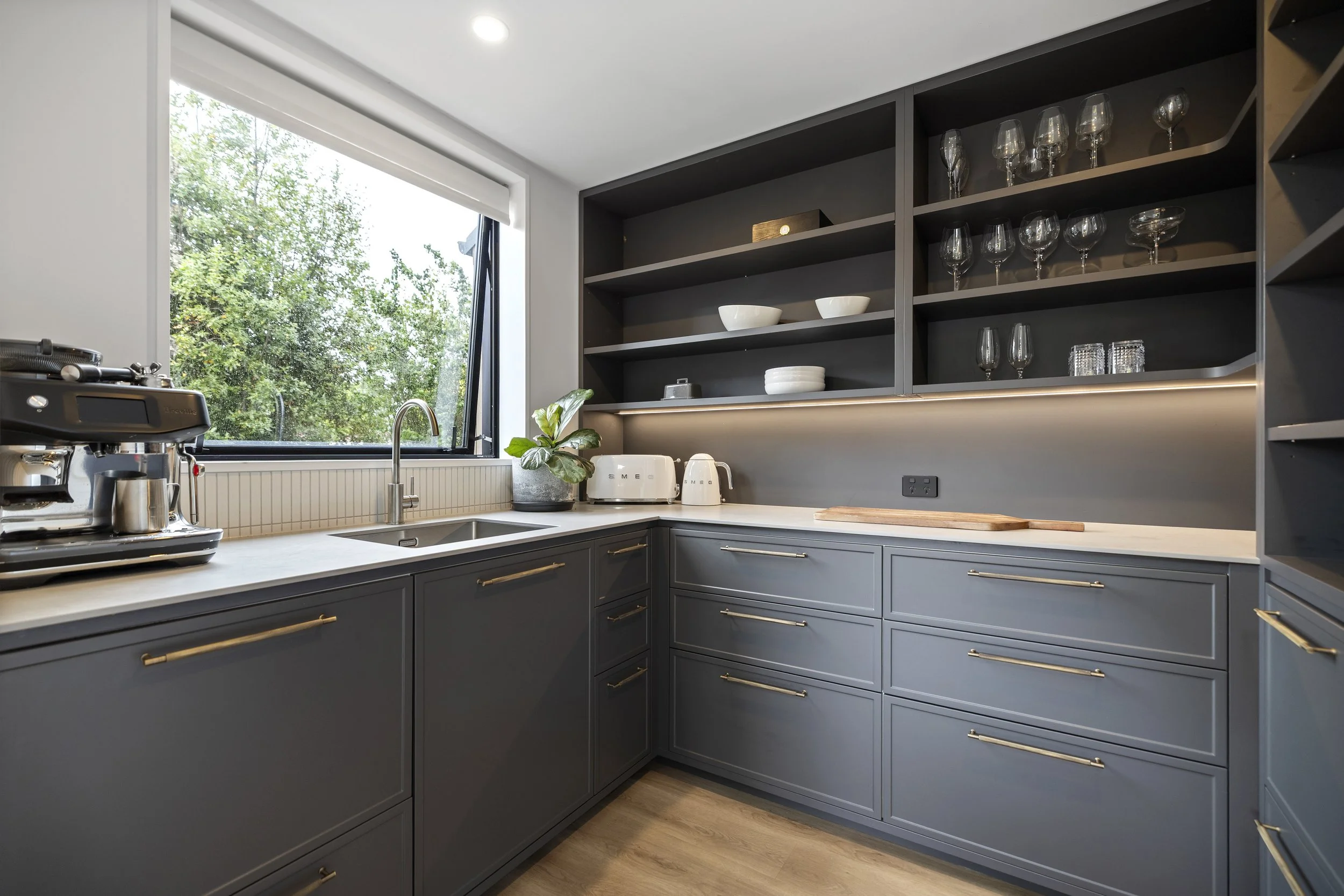 Modern kitchen with dark gray cabinets and open shelving, a large window with a view of trees, and small kitchen appliances including a toaster and espresso machine.