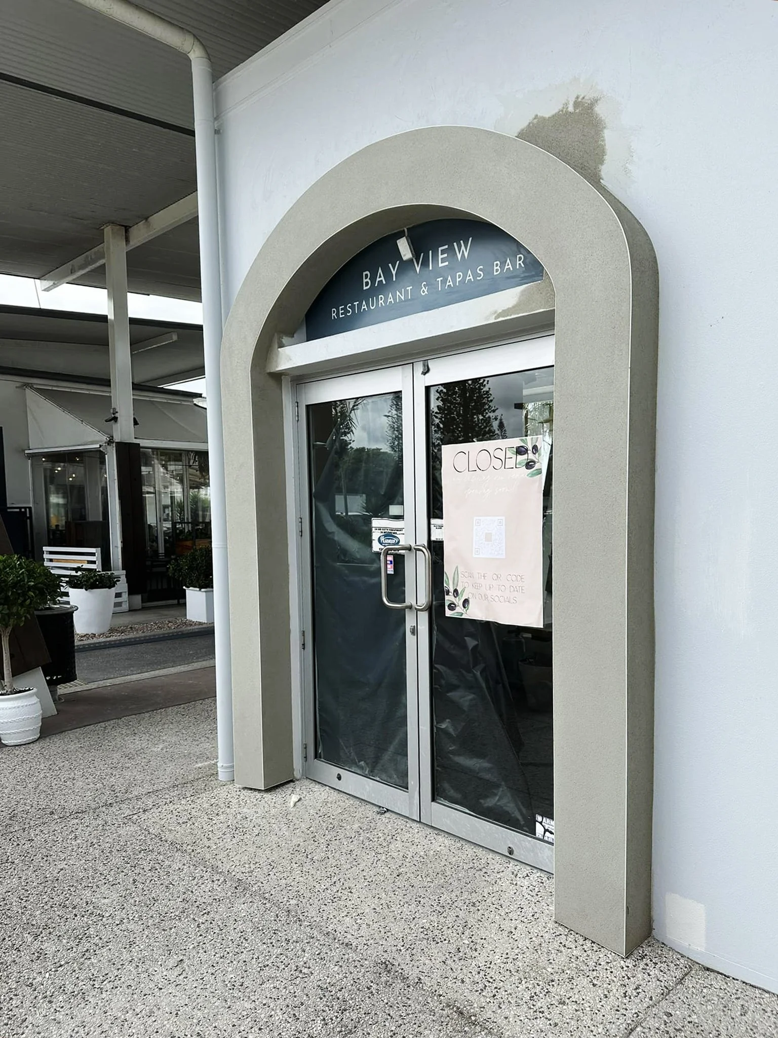 The entrance to a restaurant called Bay View with a sign on the glass door indicating it is closed. The exterior has a curved design with a textured white wall and a pinstripe ceiling.