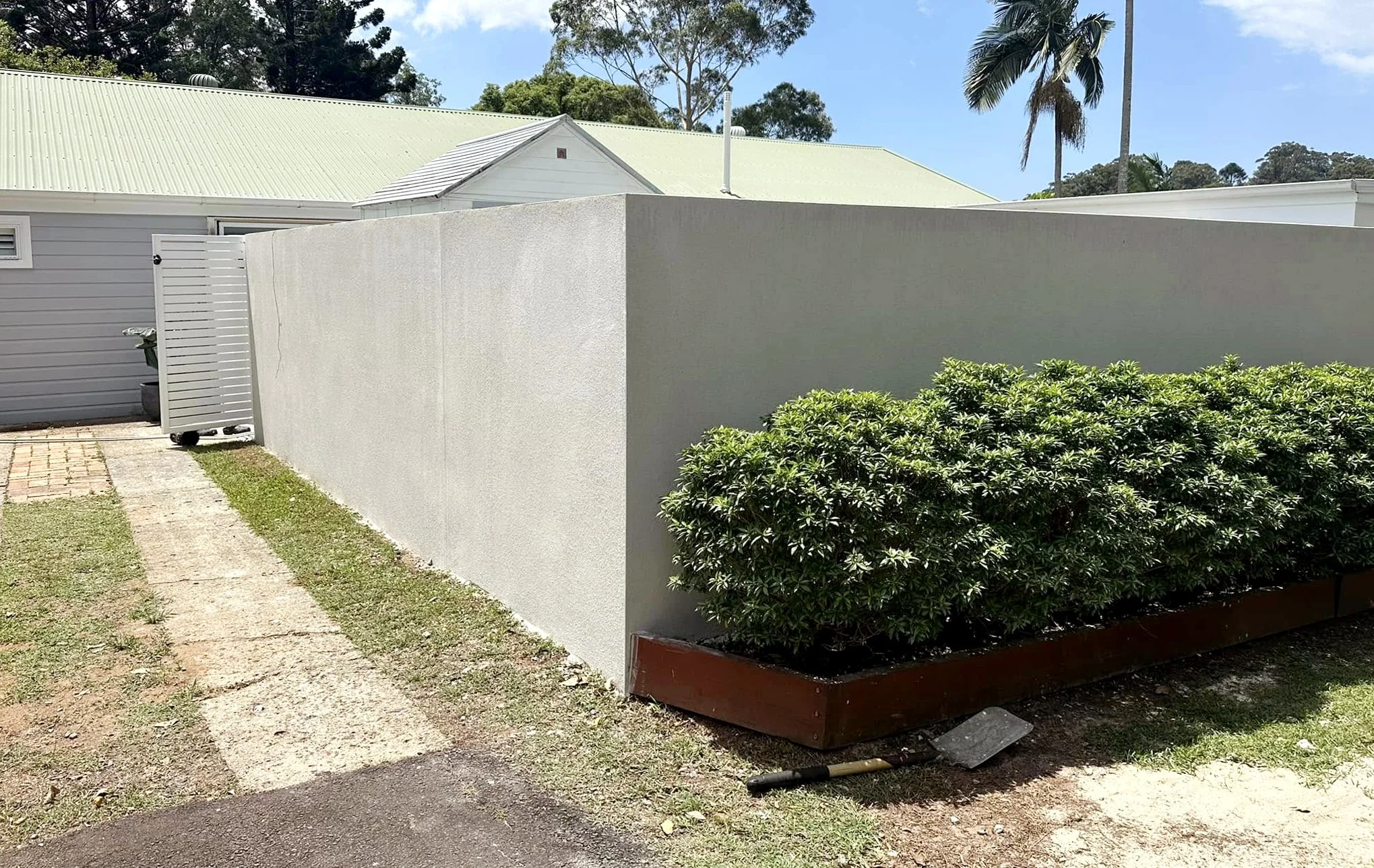 A concrete wall with a bush in a rectangular planter in front, a paved walkway with patches of grass, along with part of a small building to the left, a white gate, and a house with a light green metal roof in the background. There are trees and a cl