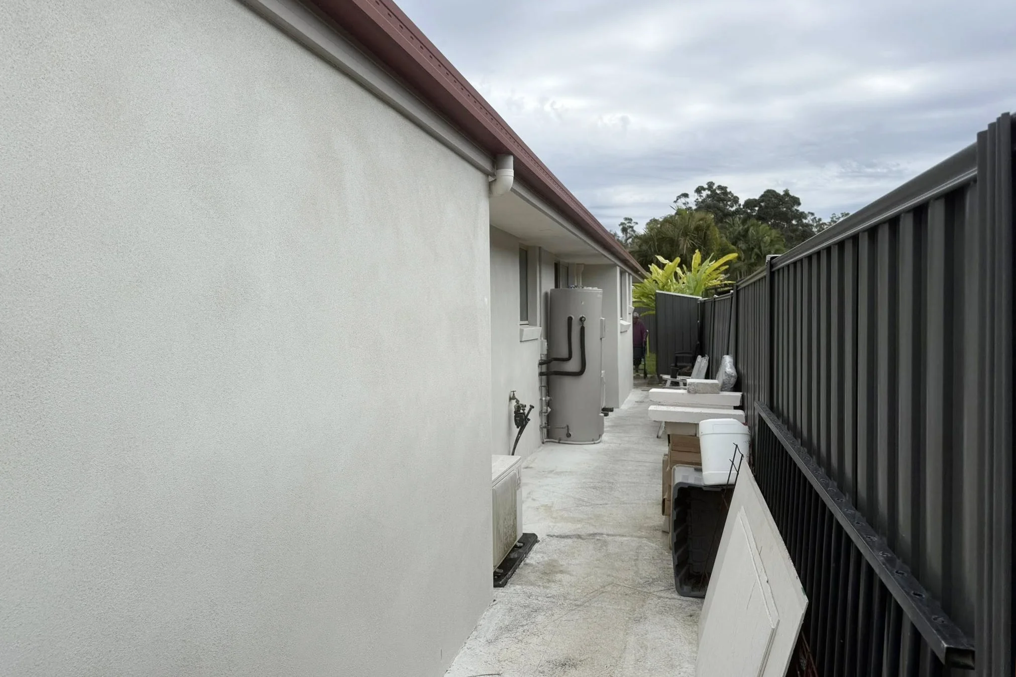 An outdoor narrow walkway between a white textured wall and a black metal fence, with various utility boxes, a white plastic bucket, and window in the background, with trees and cloudy sky beyond.