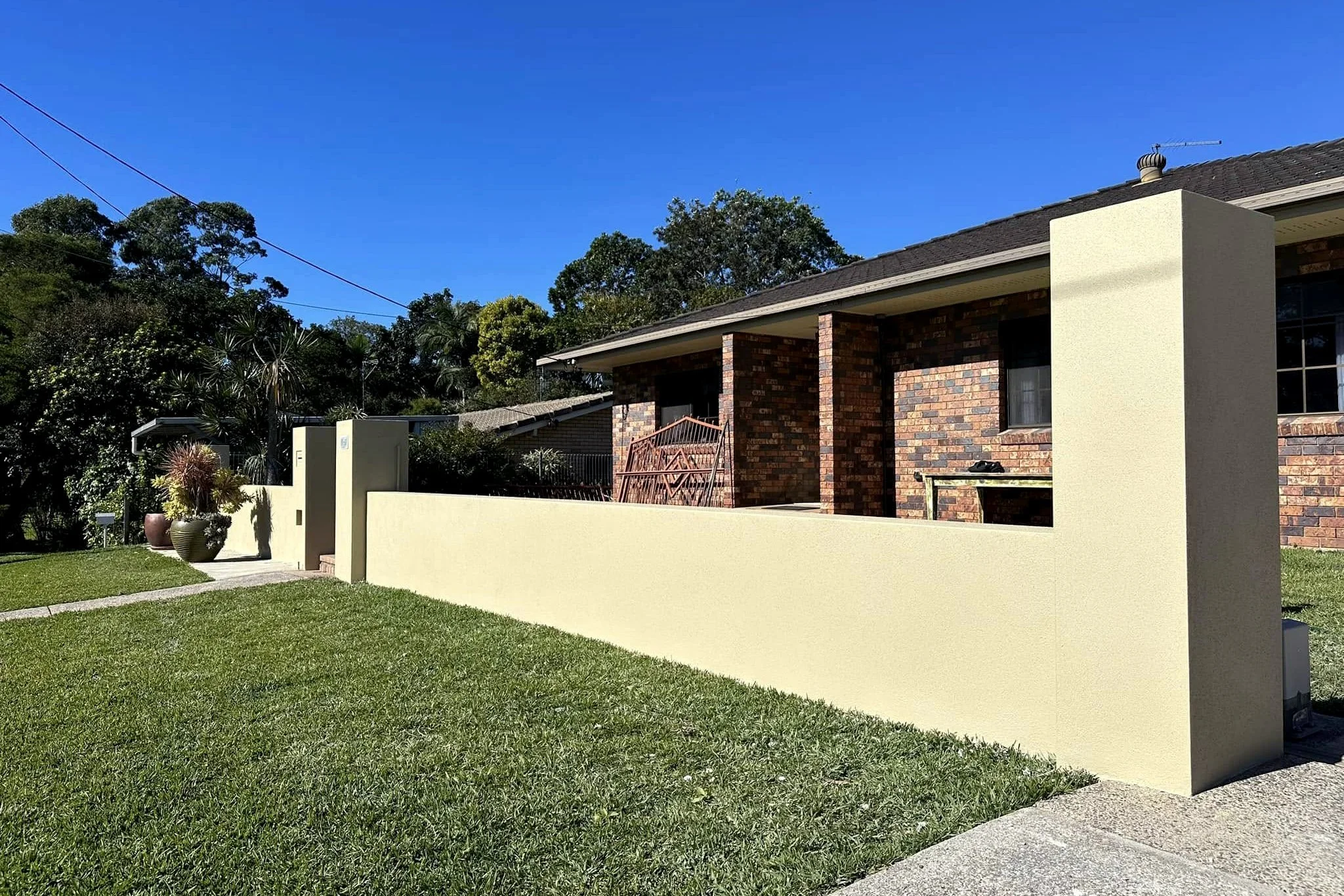 A brick house with a small porch, large windows, and a beige exterior wall on one side. There are potted plants and a well-maintained green lawn in front of the house, with tall trees and a clear blue sky in the background.