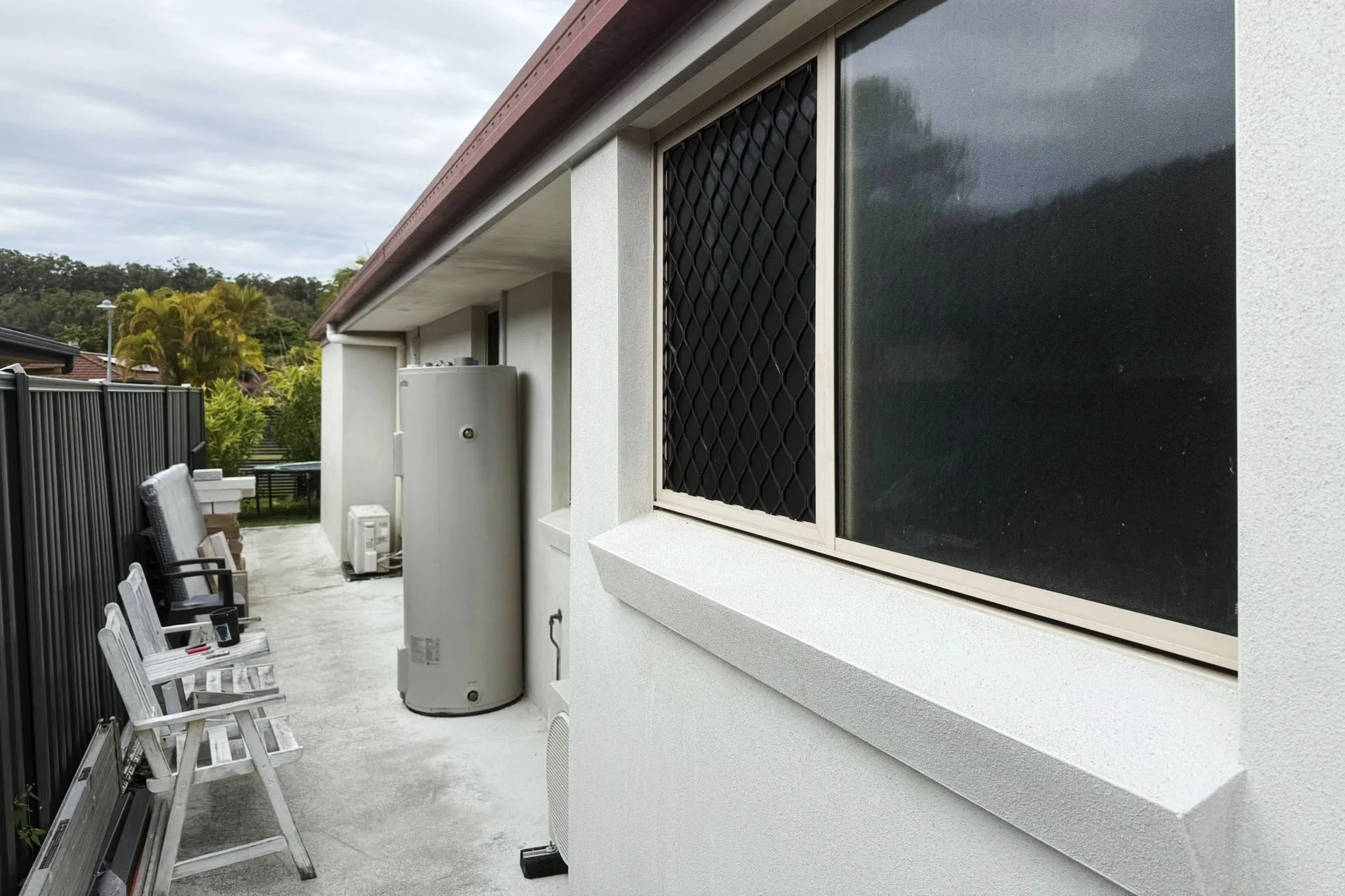 A house exterior with three chairs, a water heater, and a window with a security screen, adjacent to a black metal fence and lush green trees in the background.