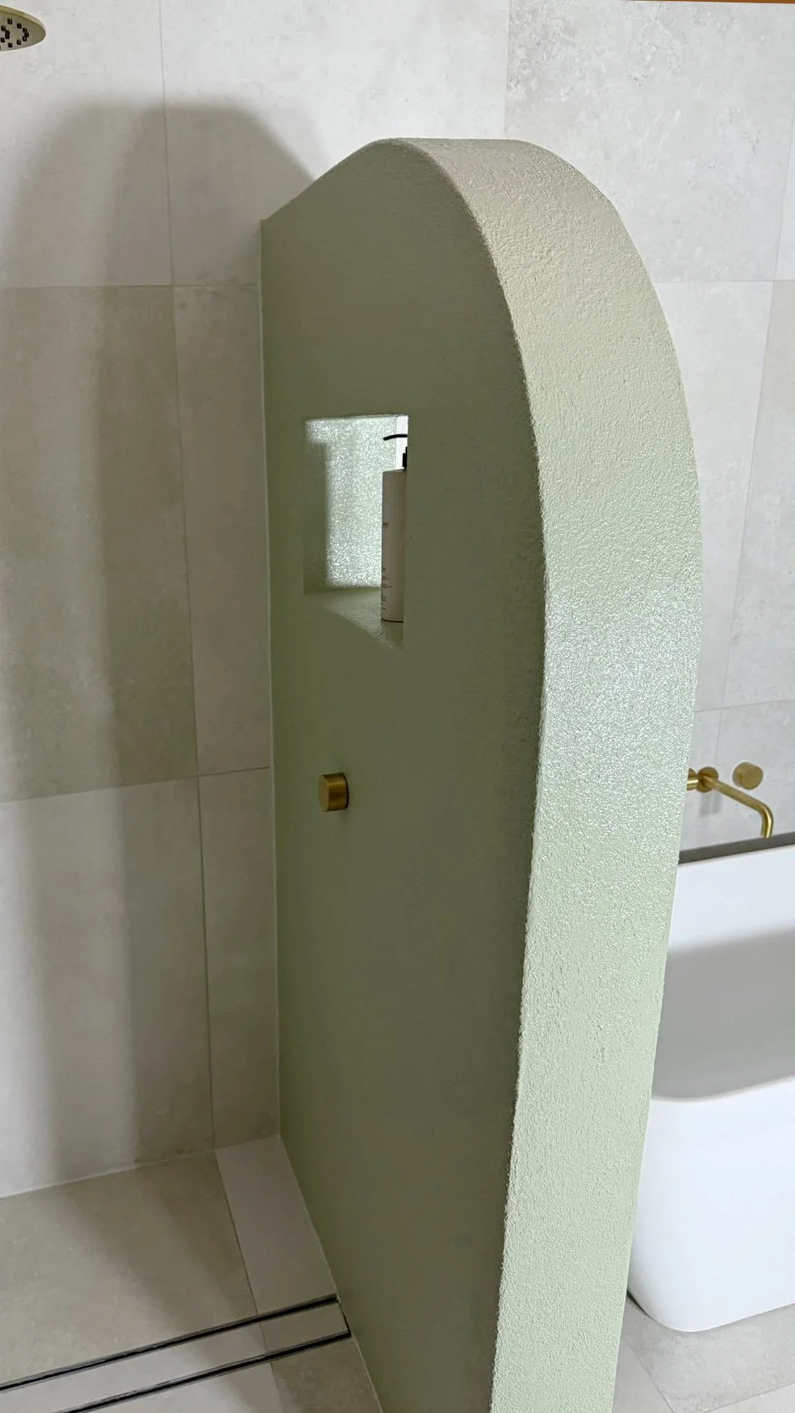 A partial view of a modern bathroom with a green textured wall, a small mirror, and a white sink with a brass faucet.