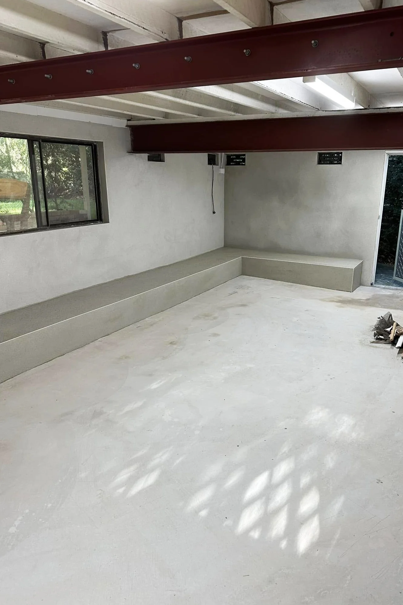 Empty unfinished room with concrete floor, white walls, a window, and exposed ceiling with beams and a red steel support beam.