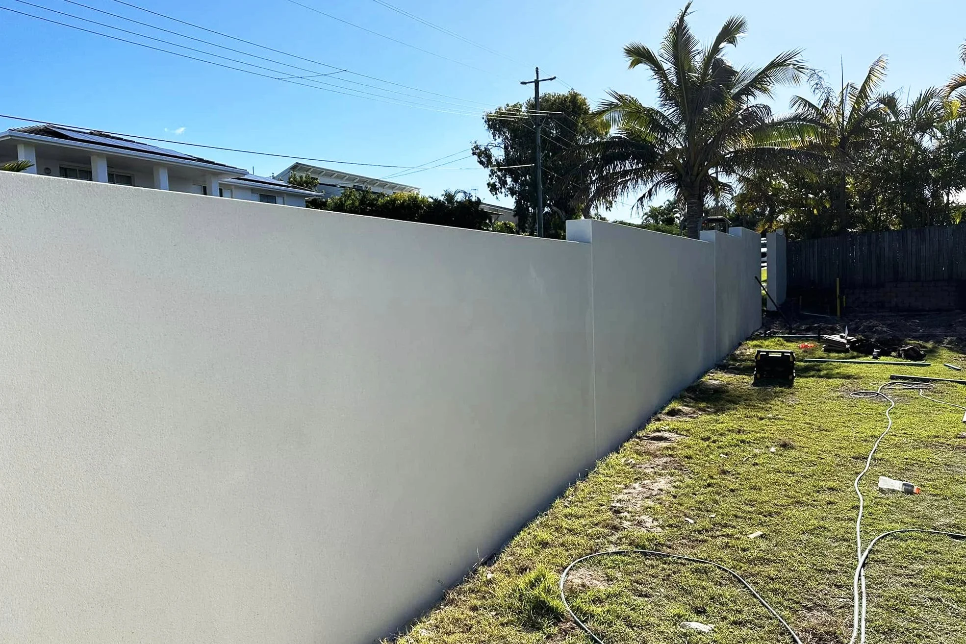 A finished white privacy wall in a backyard with trees, a sunny sky, and construction tools and hoses on the grass.