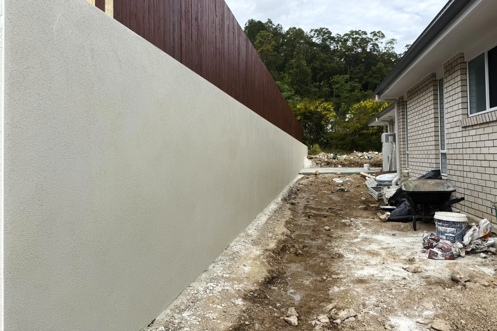 Construction site with a newly built white concrete wall on the left and a house with a brick exterior on the right, surrounded by trees in the background. The ground is uneven with dirt, rocks, and construction materials scattered around.