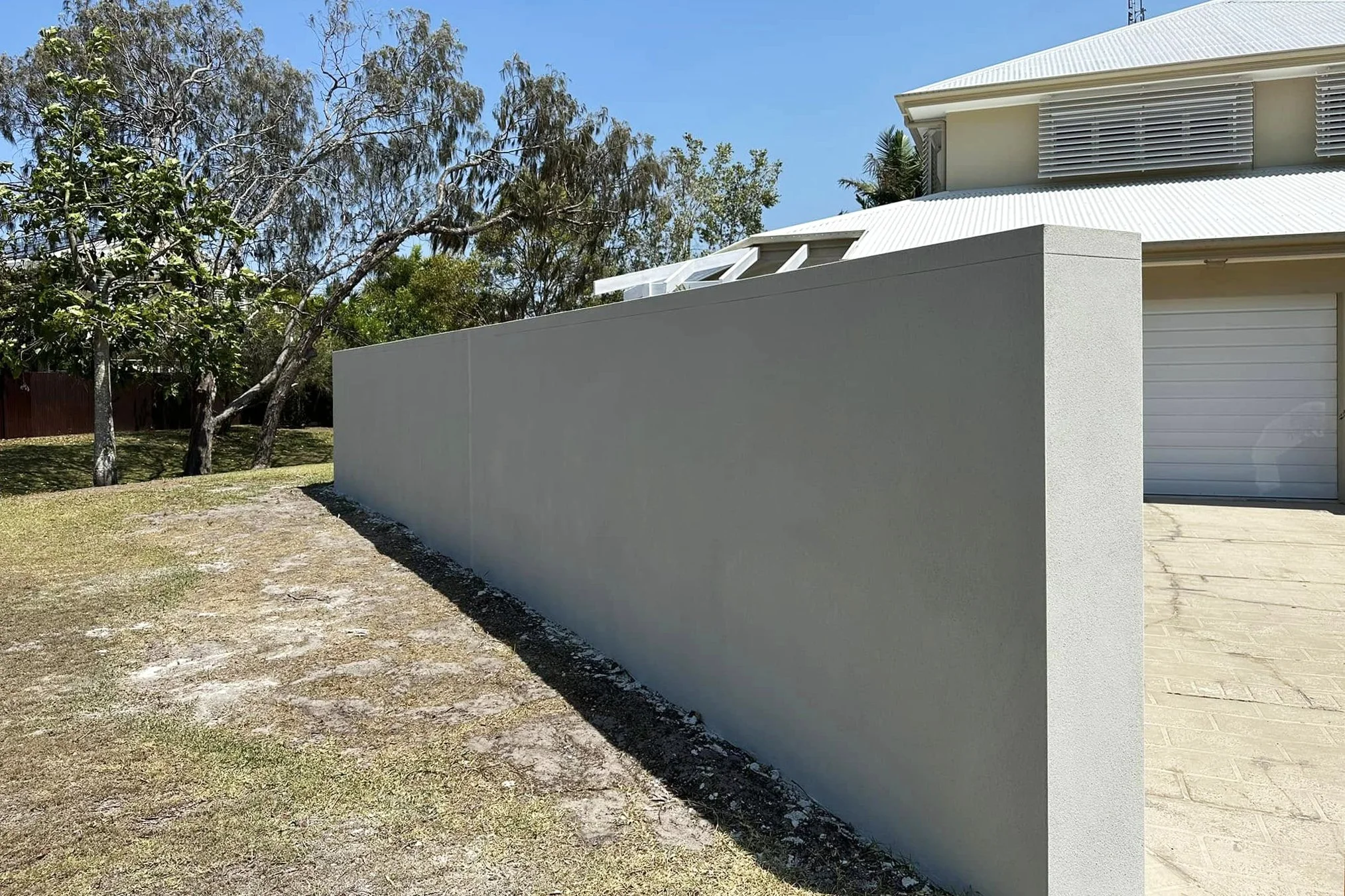 A modern house with a white metal roof, a garage door, and a gray concrete wall. There is a tree with green leaves and a blue sky in the background.
